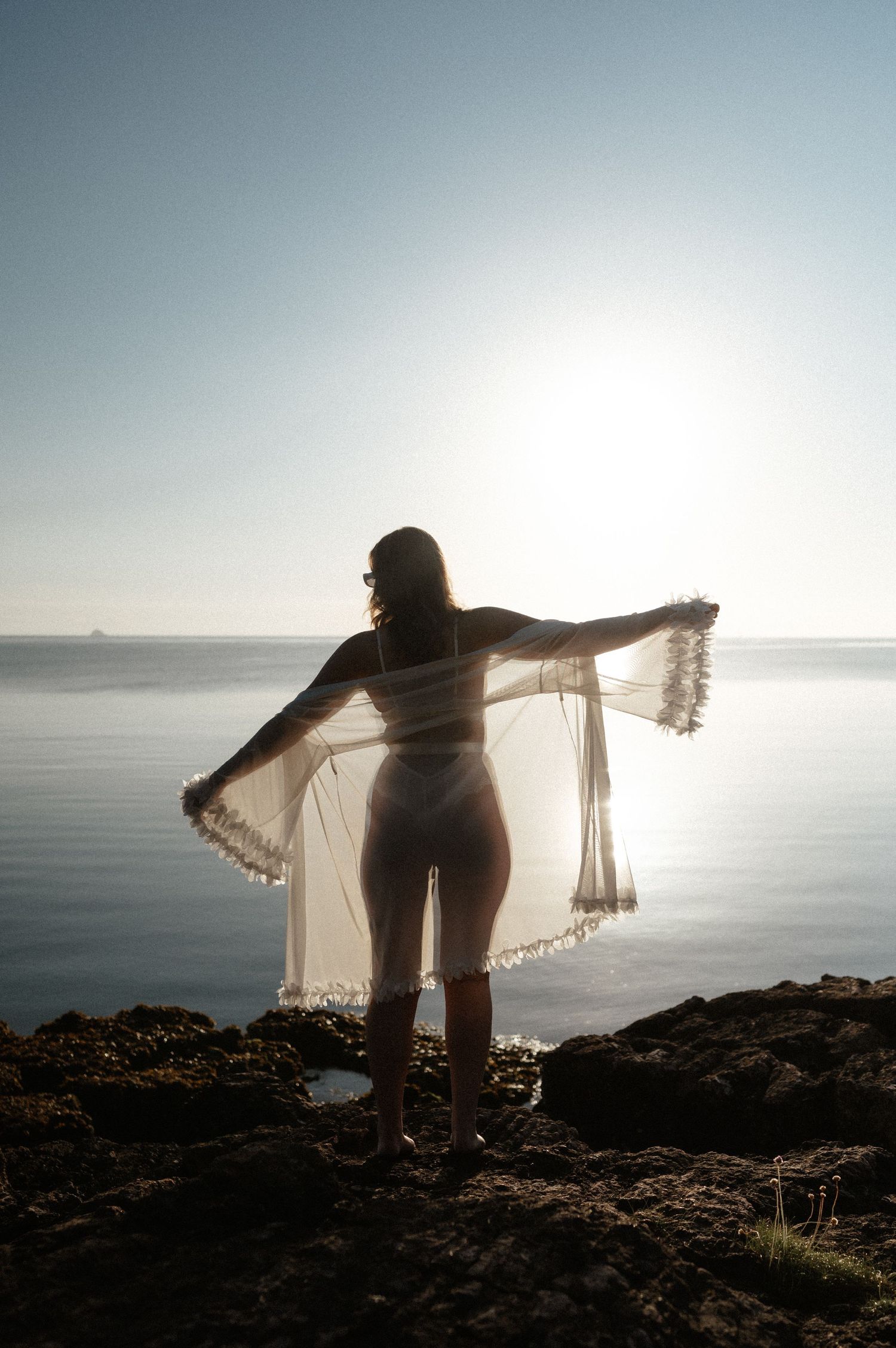 Silhouette against ocean sunset holding sheer fabric with arms outstretched.