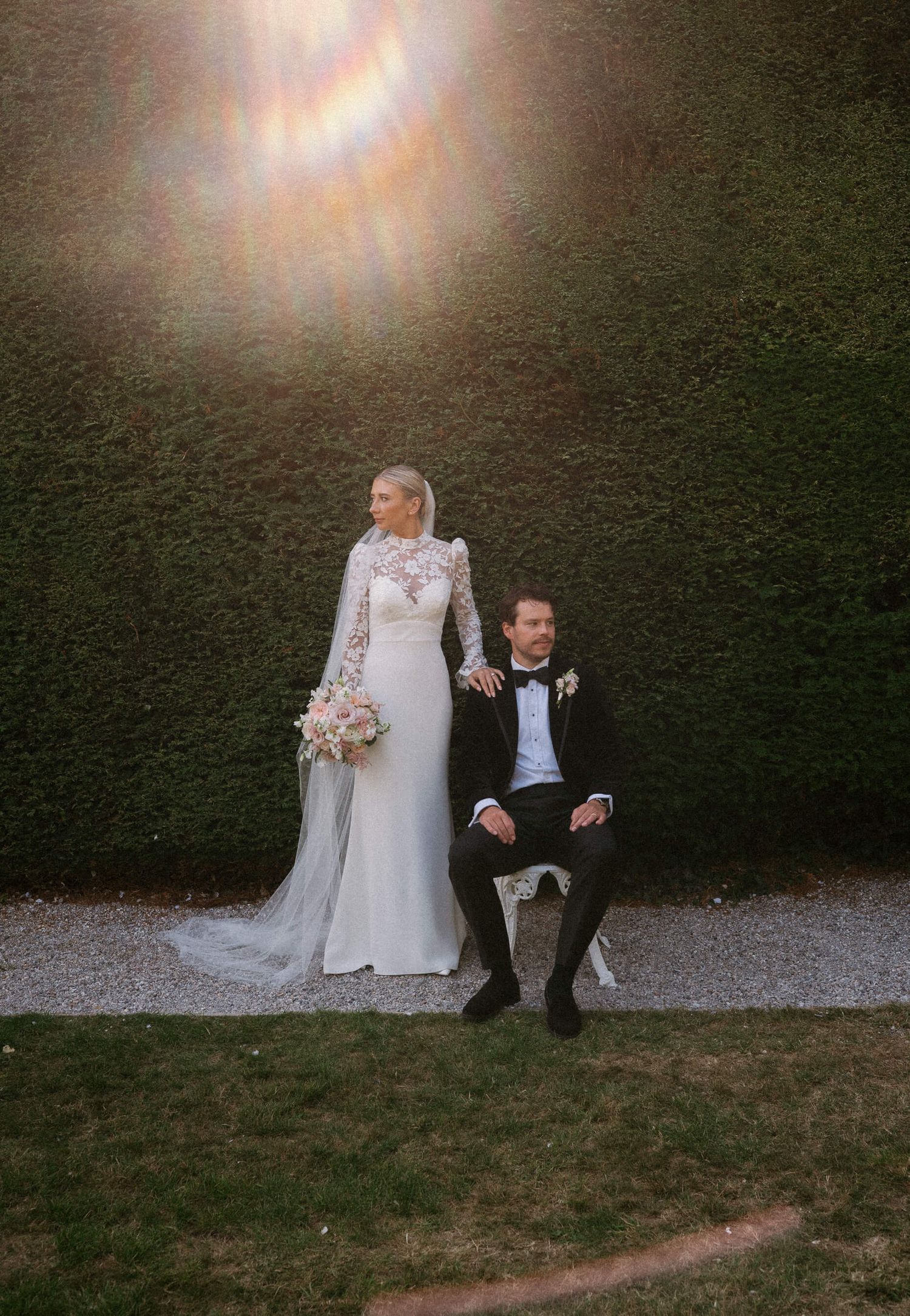 Couple in wedding attire sits on grass with dramatic sunburst lighting effect.
