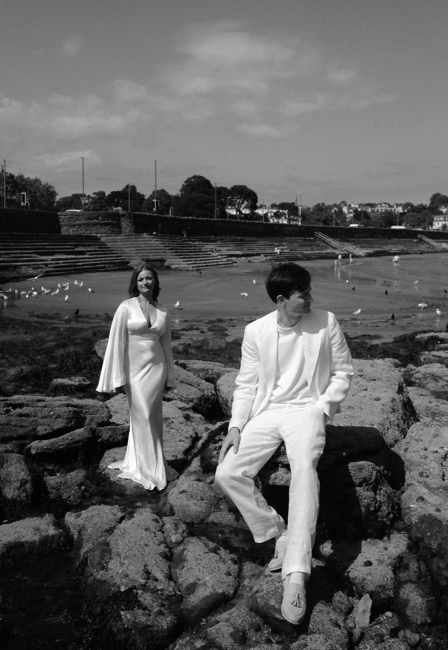 People in white outfits sitting on rocky coastal formations with ocean and buildings visible in background.