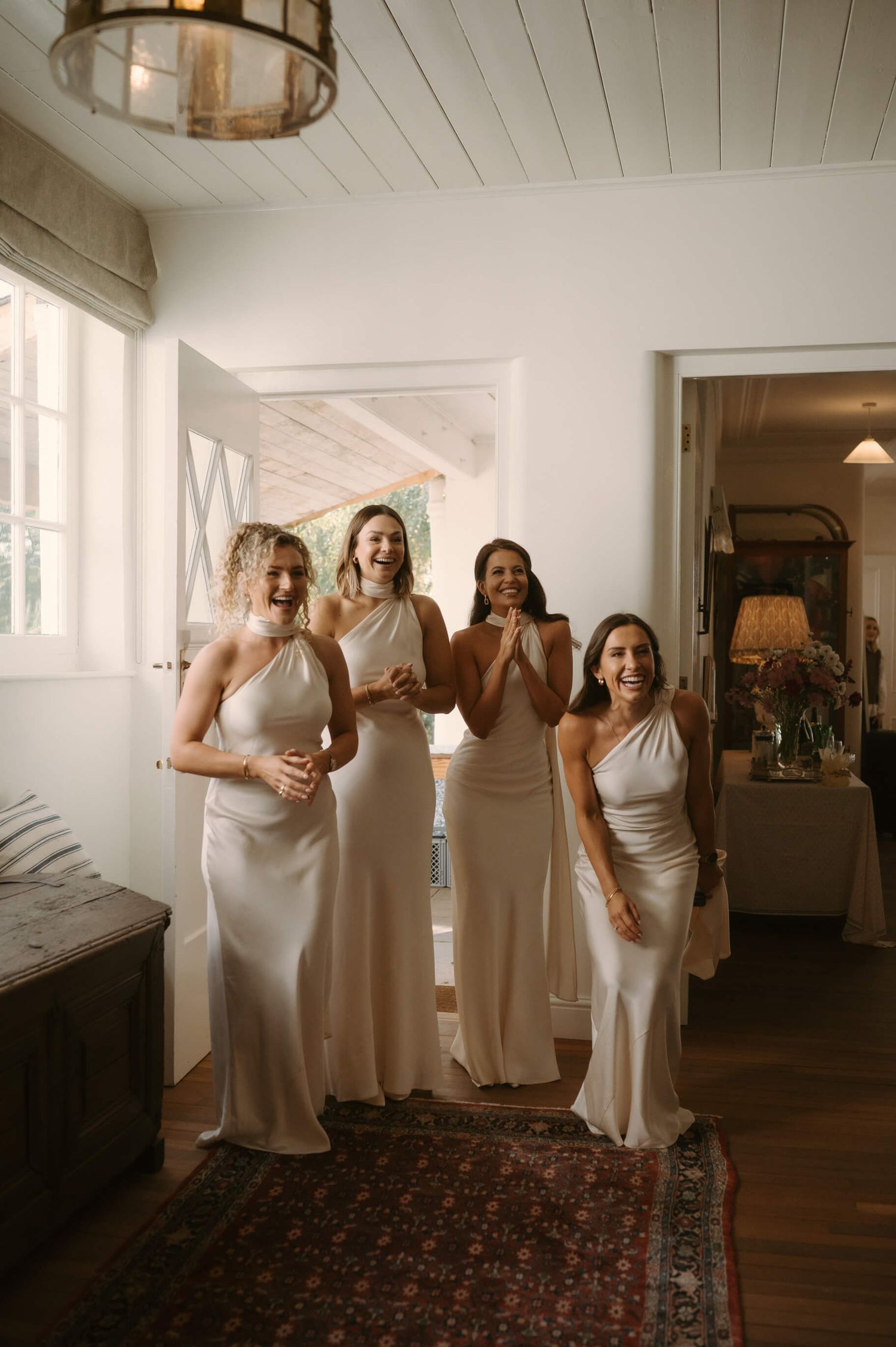Bridesmaids in white silk dresses share an excited moment in a bright hallway.