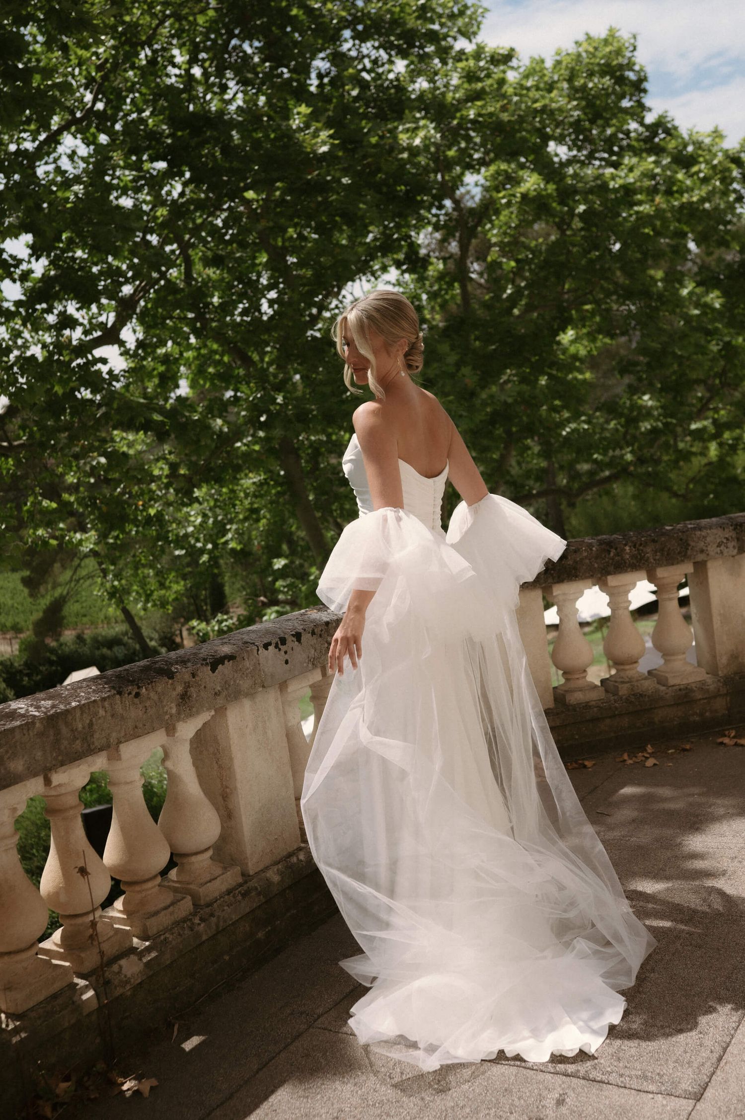 Flowing white wedding dress photographed on a stone balcony surrounded by lush greenery.