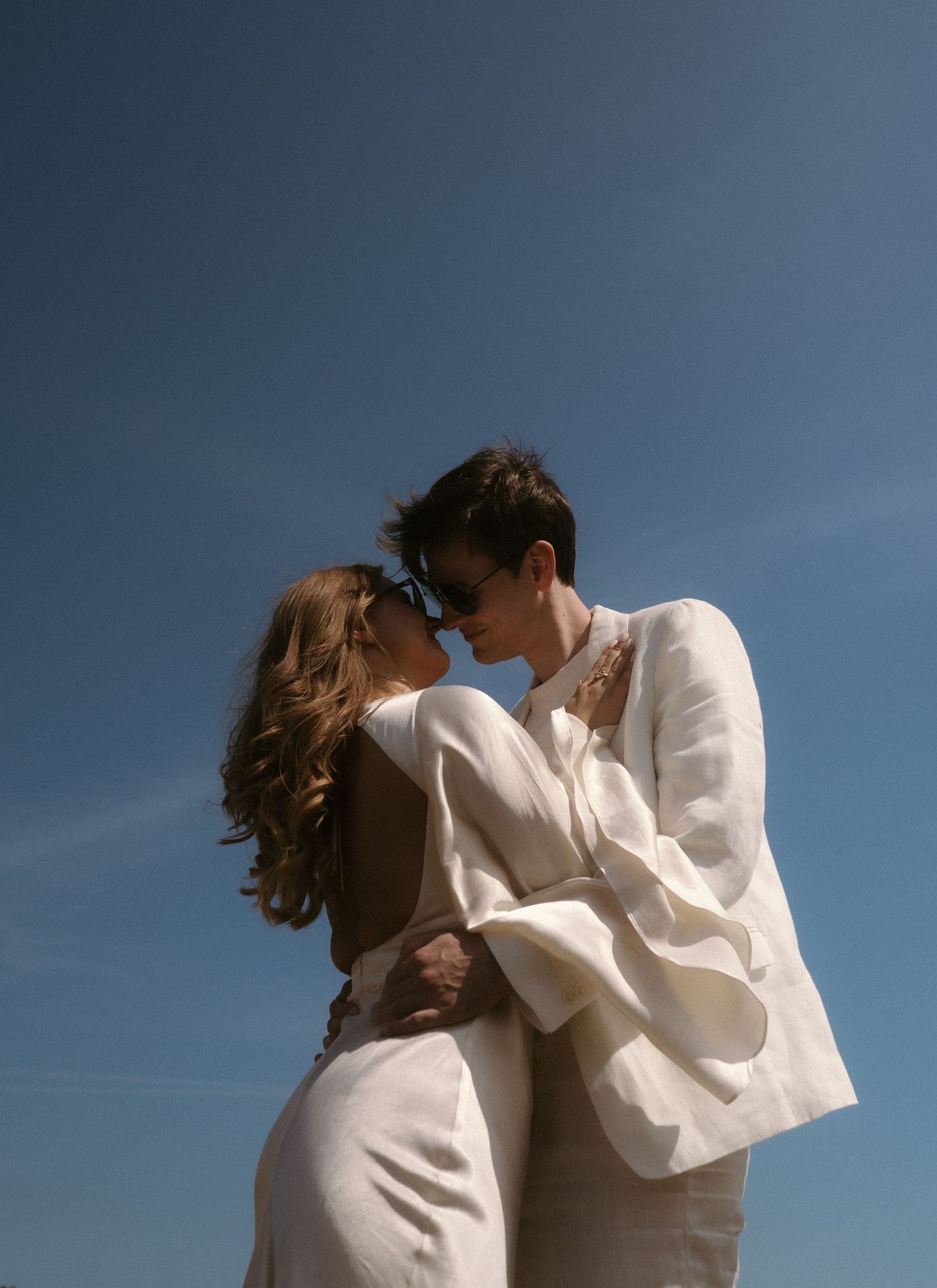 A couple wearing white outfits embraces romantically against a blue sky background in a series of candid moments.