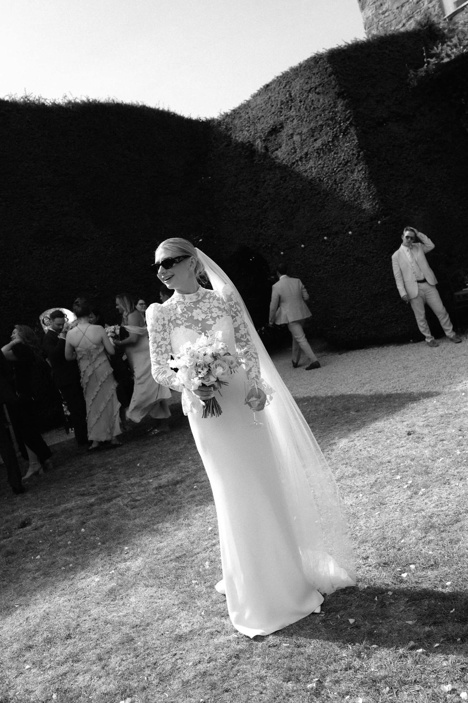 Black and white photo of a bride in a sleek dress and veil holding a bouquet outdoors.