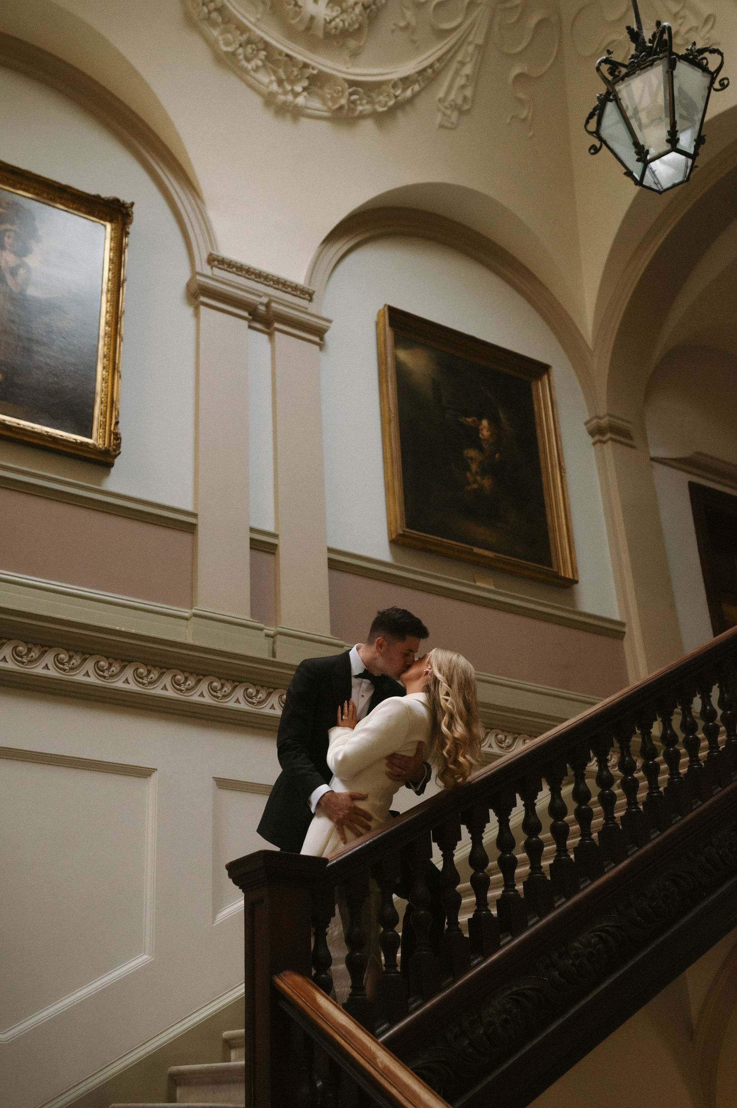 A couple shares a romantic kiss on an elegant staircase in a historic building with ornate decor and paintings.