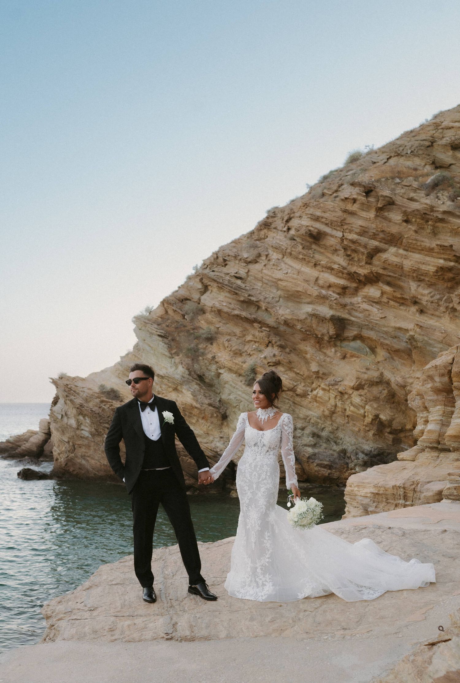 A couple poses along dramatic coastal cliffs with white rocky formations and blue Mediterranean waters.