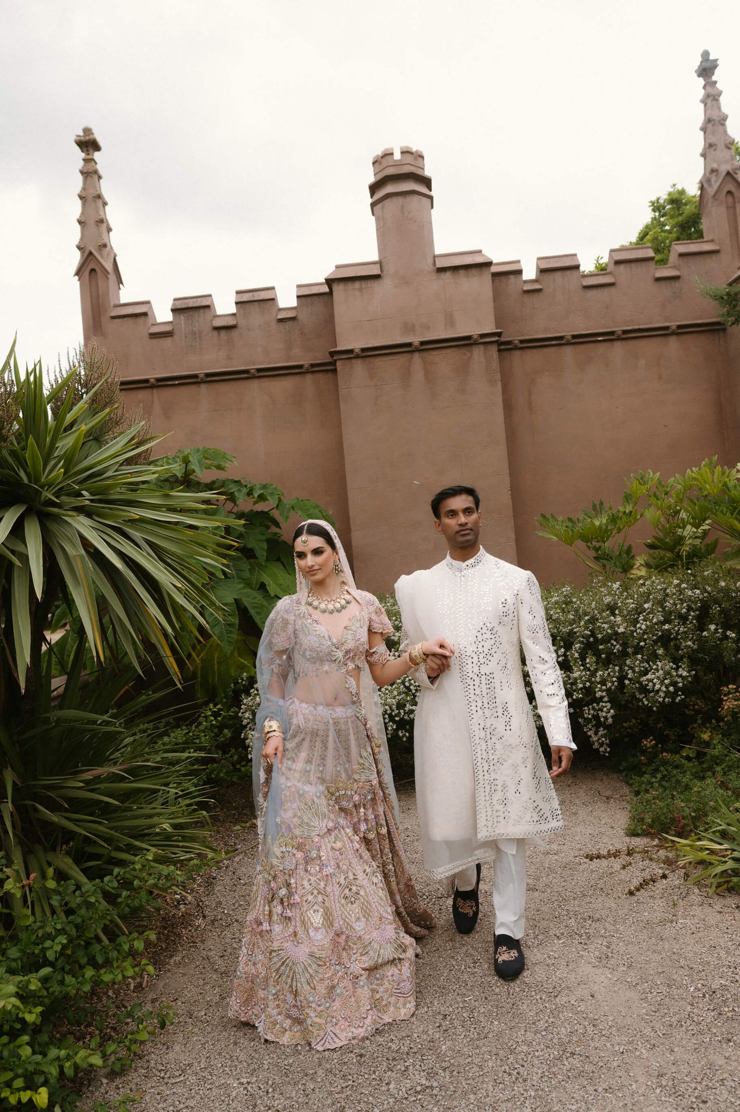 Couple in traditional South Asian wedding attire walking along a castle-like path with palm trees.