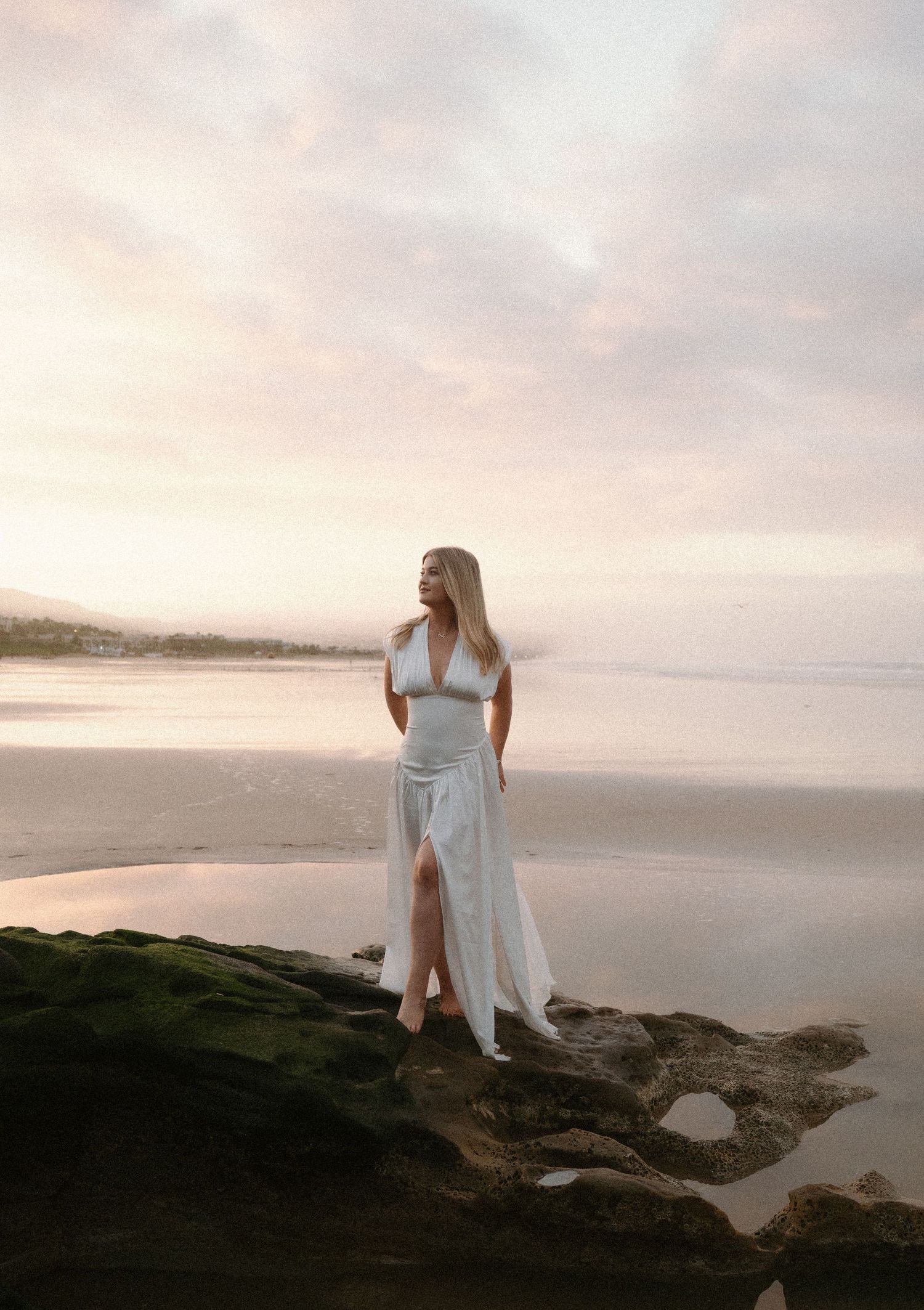 A person in a white dress stands on coastal rocks during sunset at a beach with soft pink skies in the background.