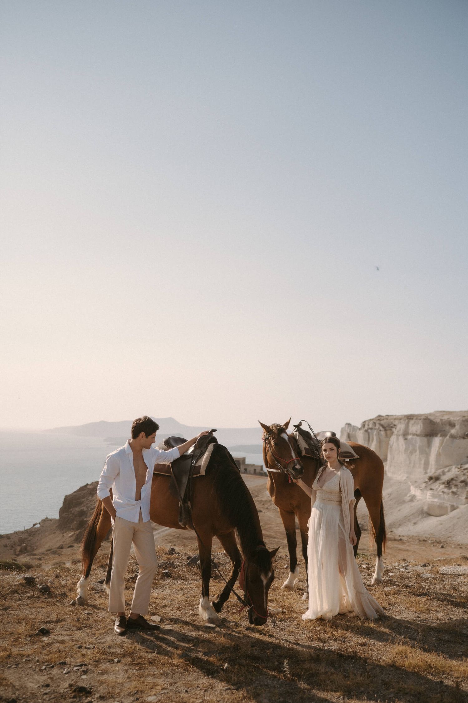 Wedding party poses with horses against dramatic cliffside backdrop at sunset.