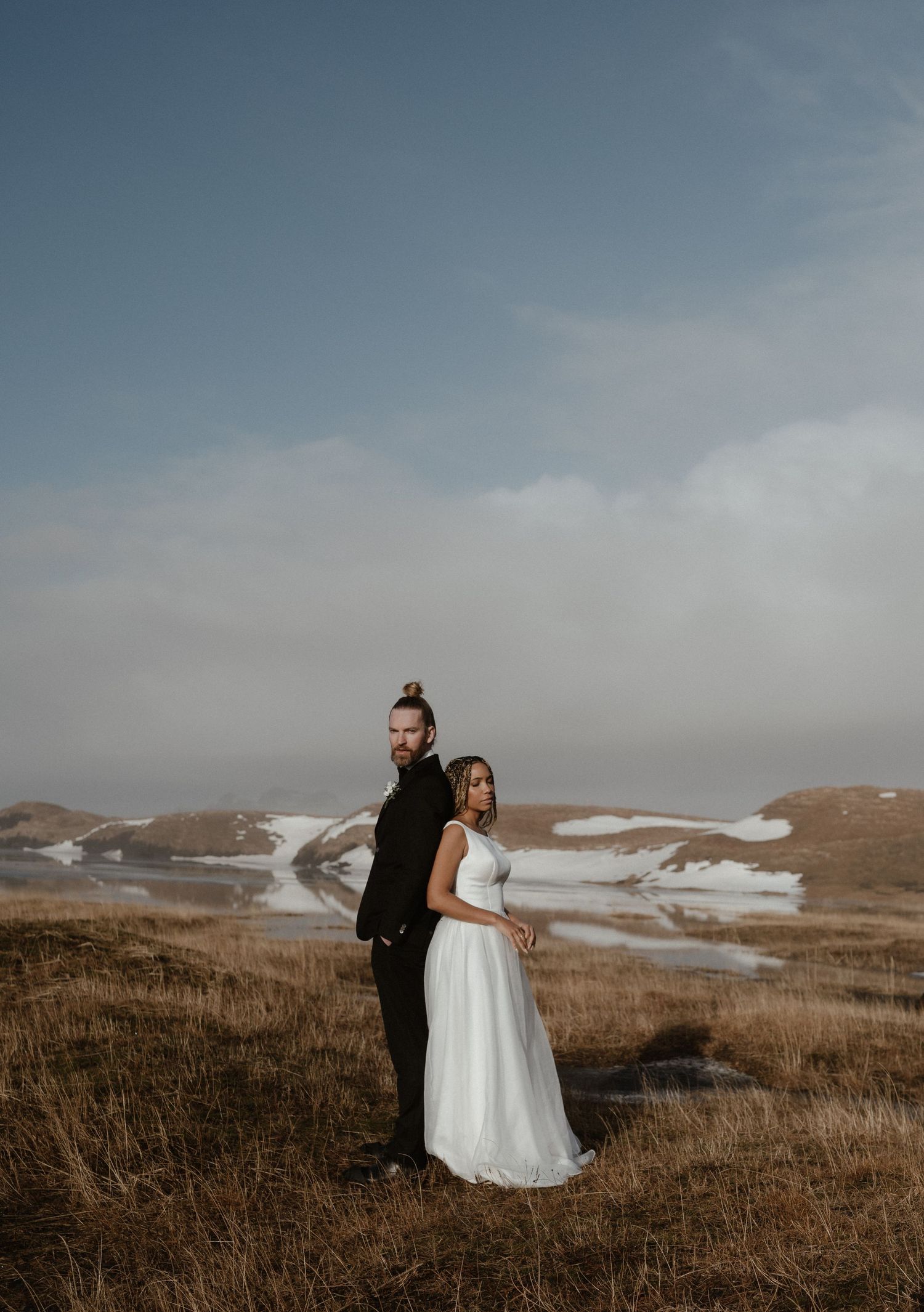 A couple in wedding attire stands together in a scenic mountain landscape with patches of snow and dry grass.