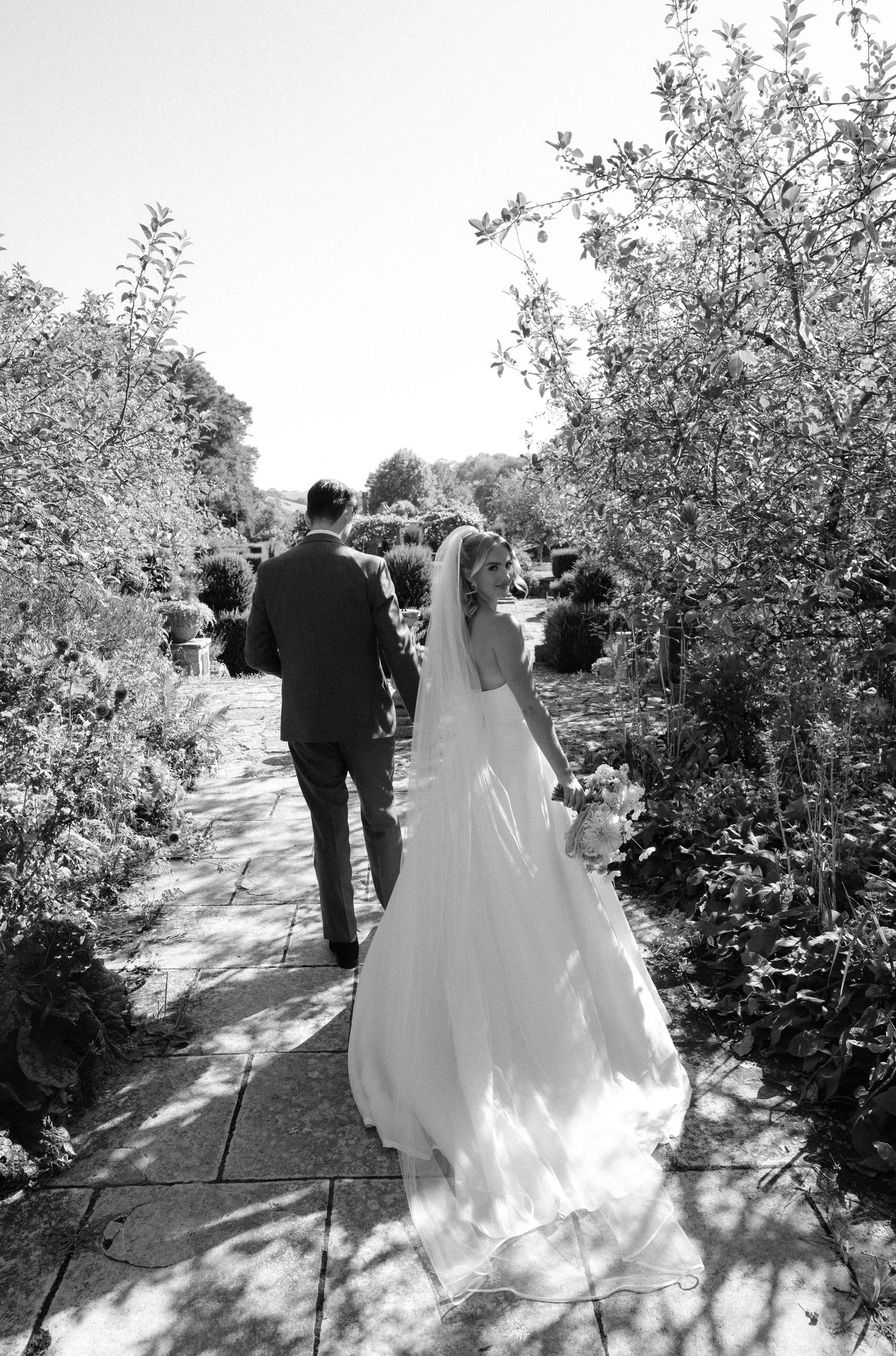 Bride in a long white gown and veil walks hand in hand with the groom along a sunlit garden path lined with trees in black and white.