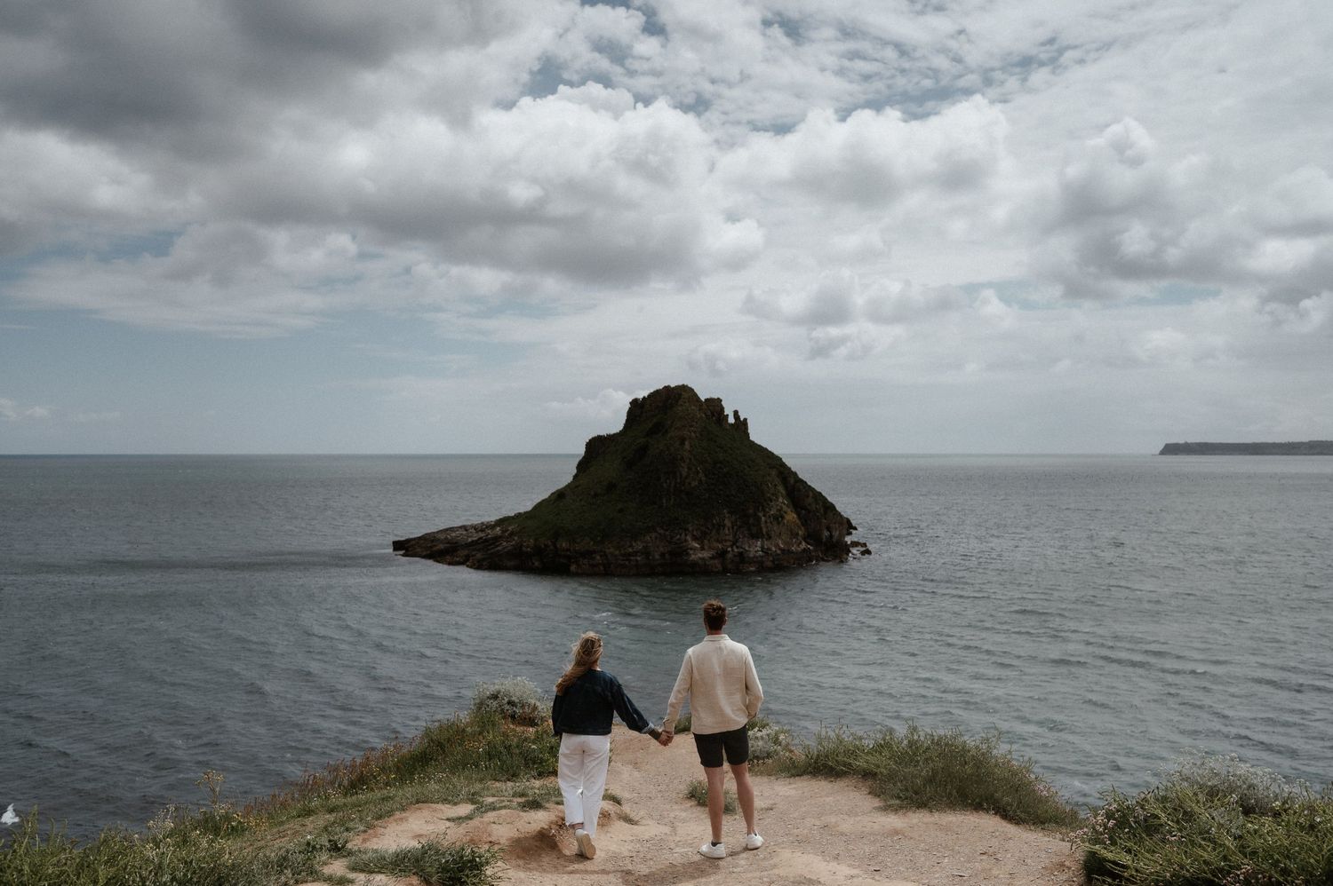 Two people stand on a coastal cliff overlooking a dramatic rocky island formation in the ocean.