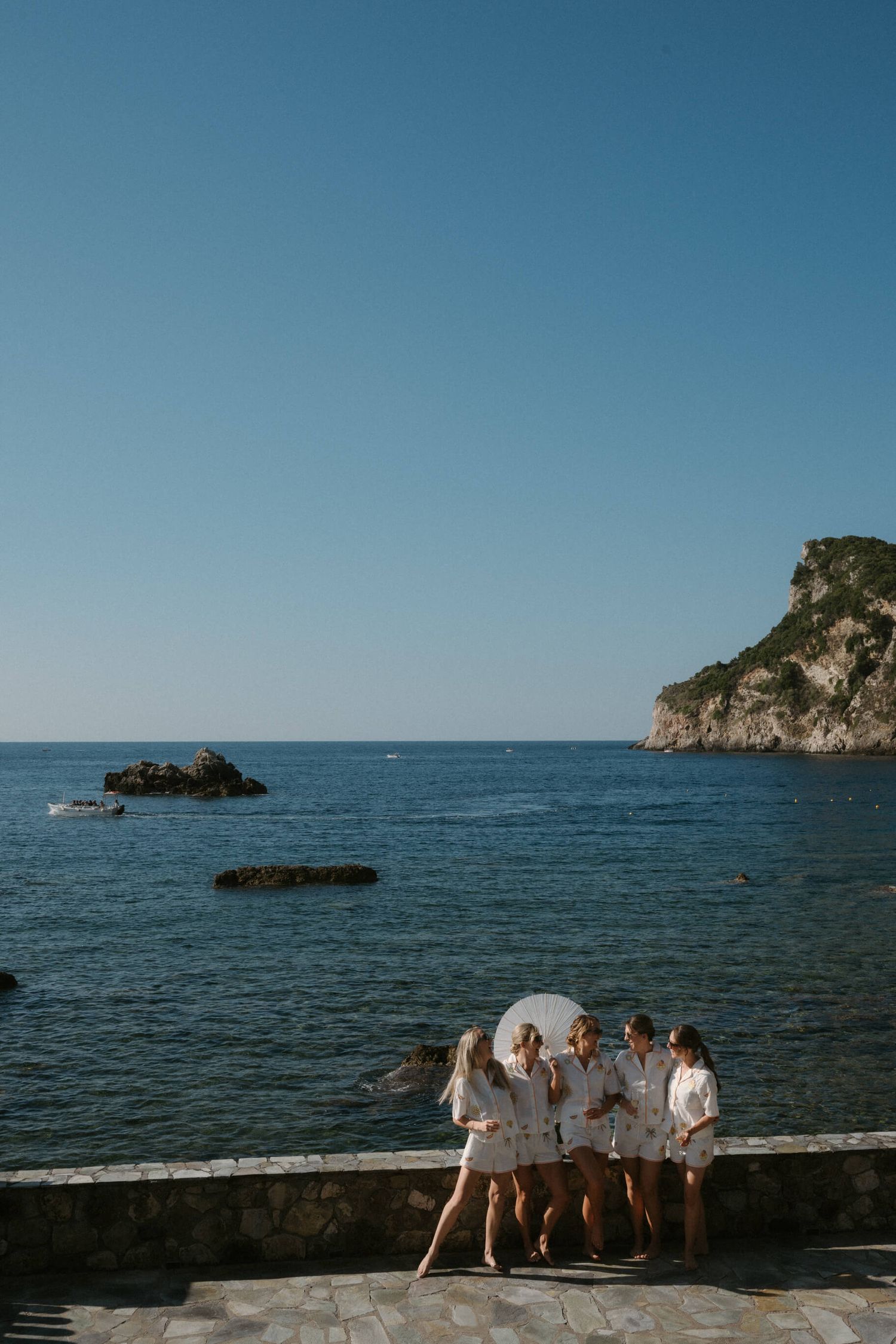 Wedding party in white attire poses along rocky coastline with blue ocean backdrop.