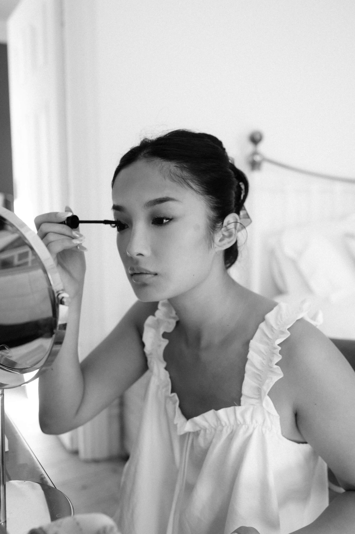 Black and white portrait of someone applying mascara in front of a mirror while wearing a white ruffled top.
