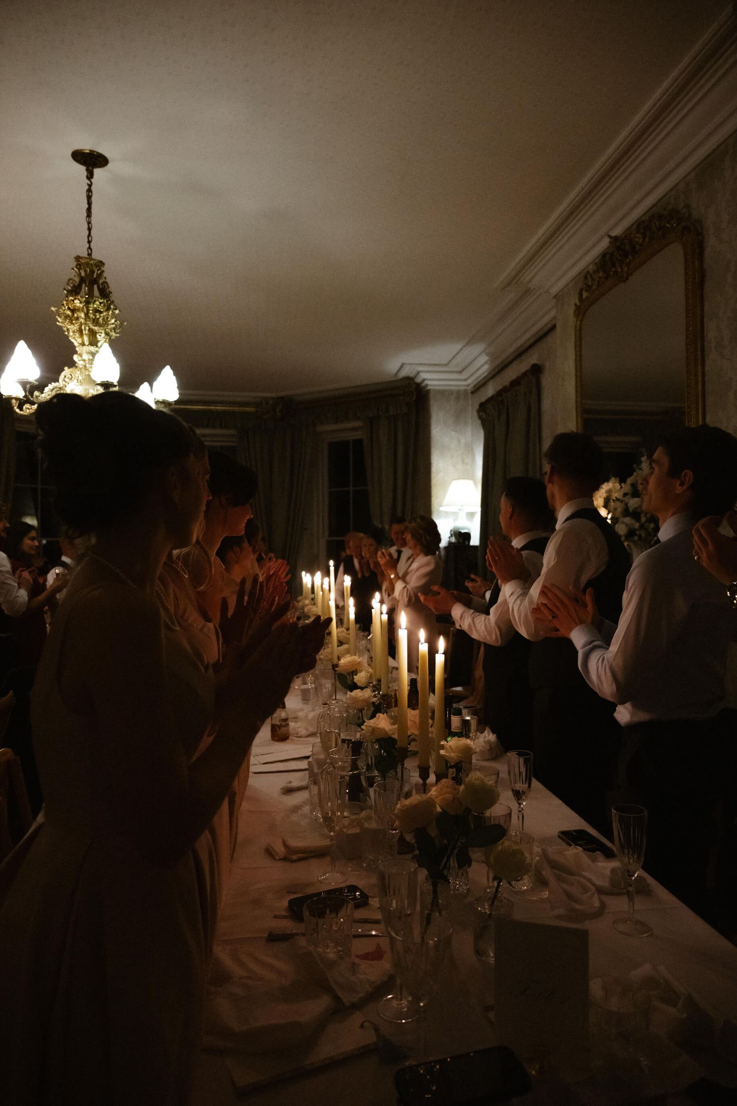 Wedding guests applaud at a candlelit dinner reception in an elegant dining room.