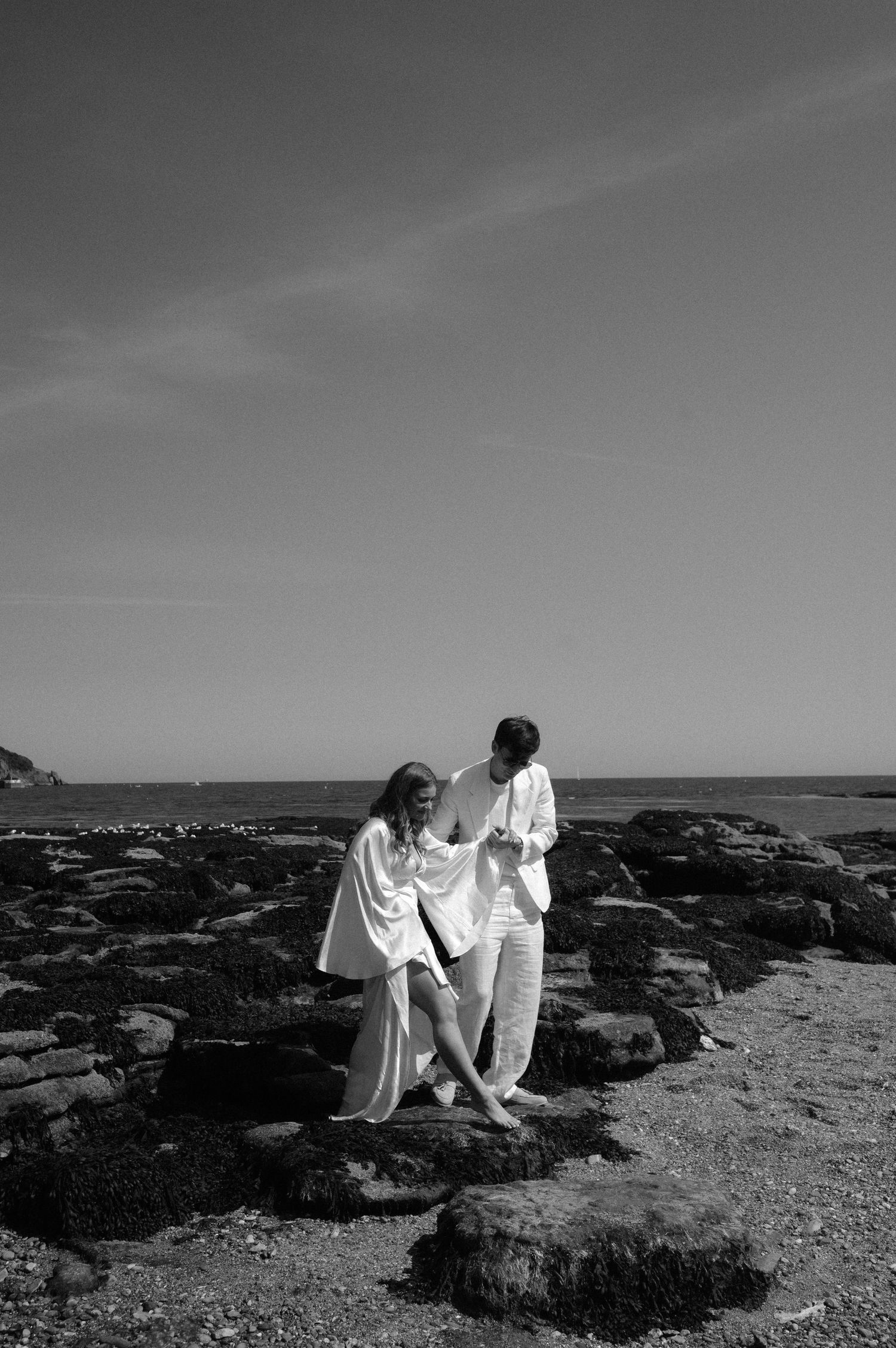 Black and white photo of couple in white clothing on rocky beach landscape.