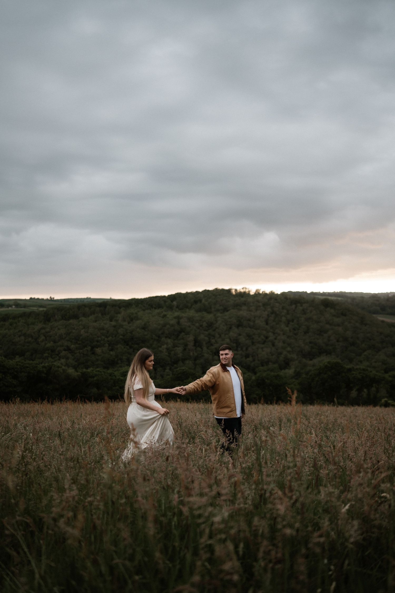 A couple poses in a moody field during sunset with dramatic clouds overhead.