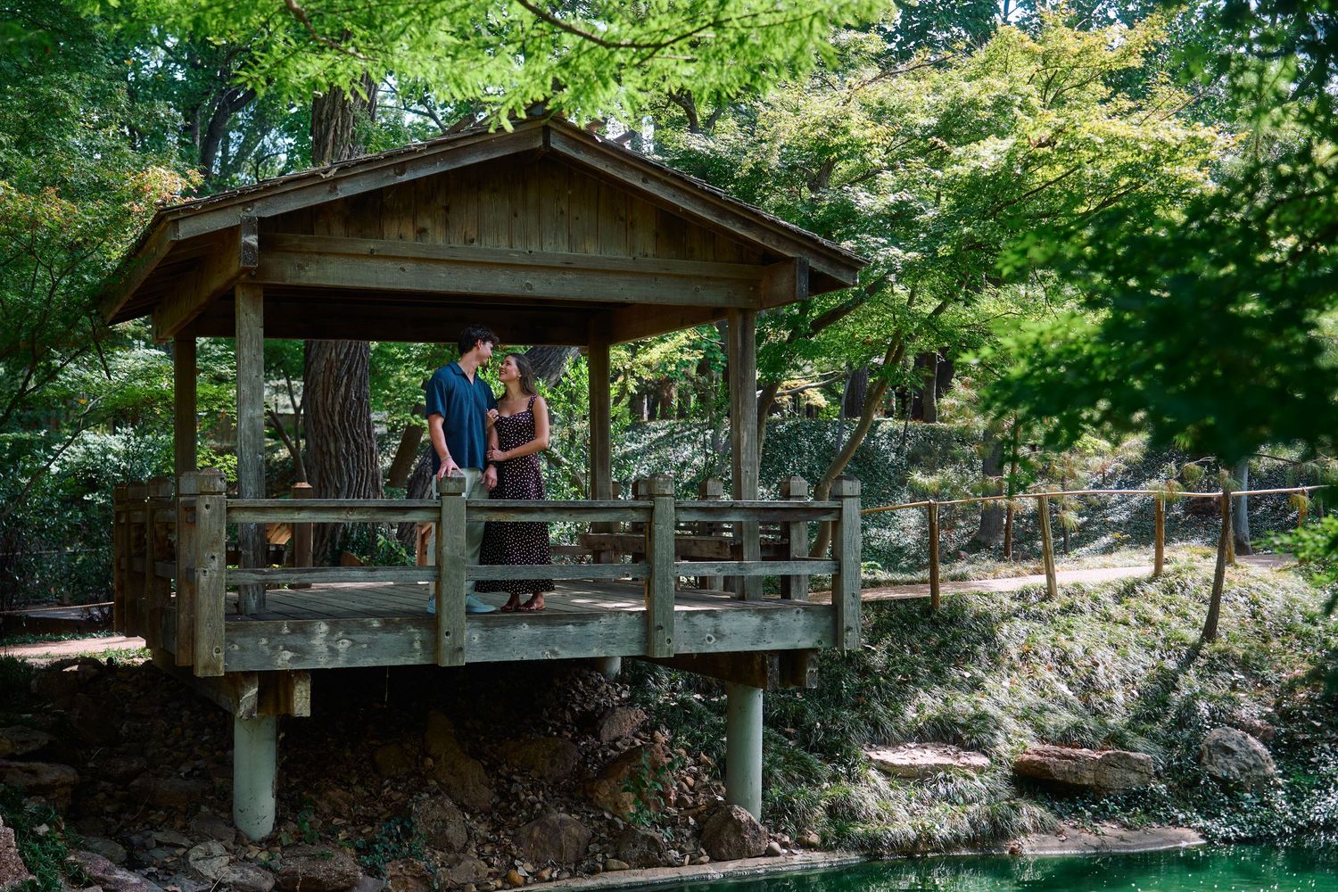 Couple standing together in a rustic wooden gazebo surrounded by lush green trees in a serene garden setting.