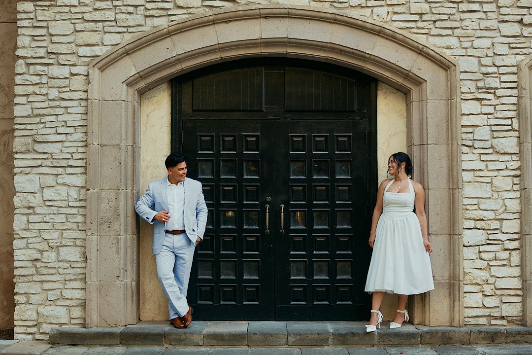 Wedding party members stand outside a stone church with dark green doors on a sunny day.