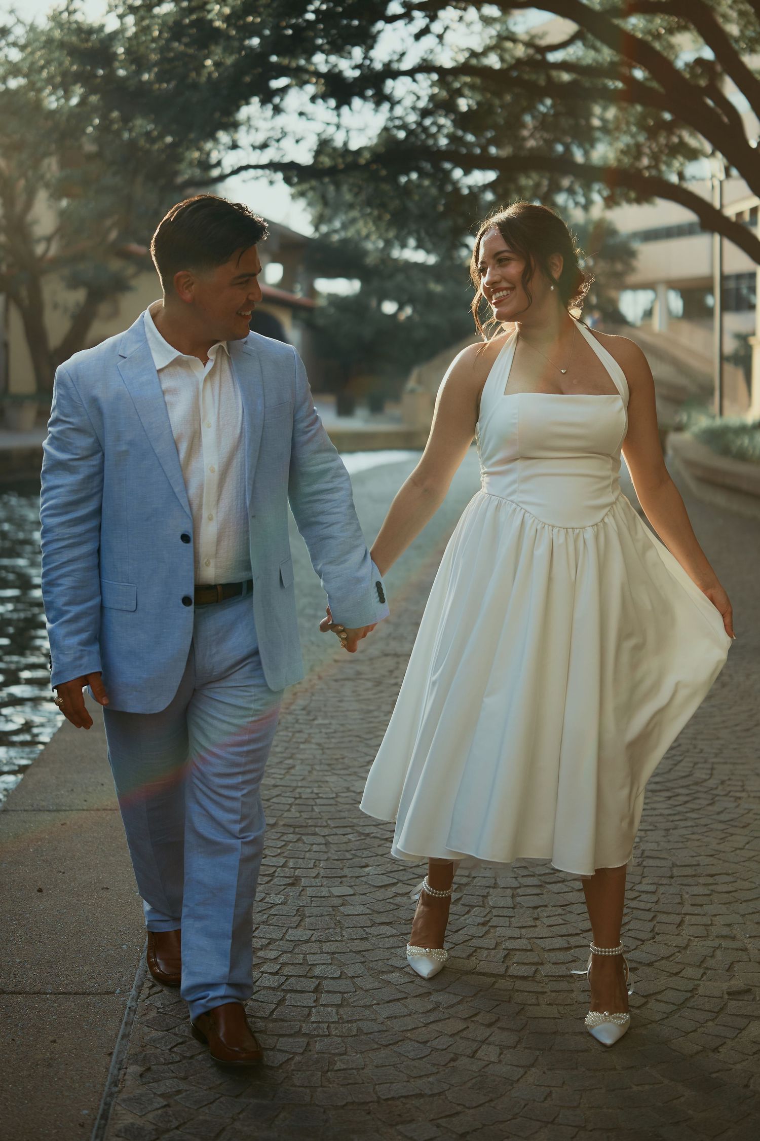 Couple walking together on cobblestone path at sunset, with woman in white dress and man in light blue suit.