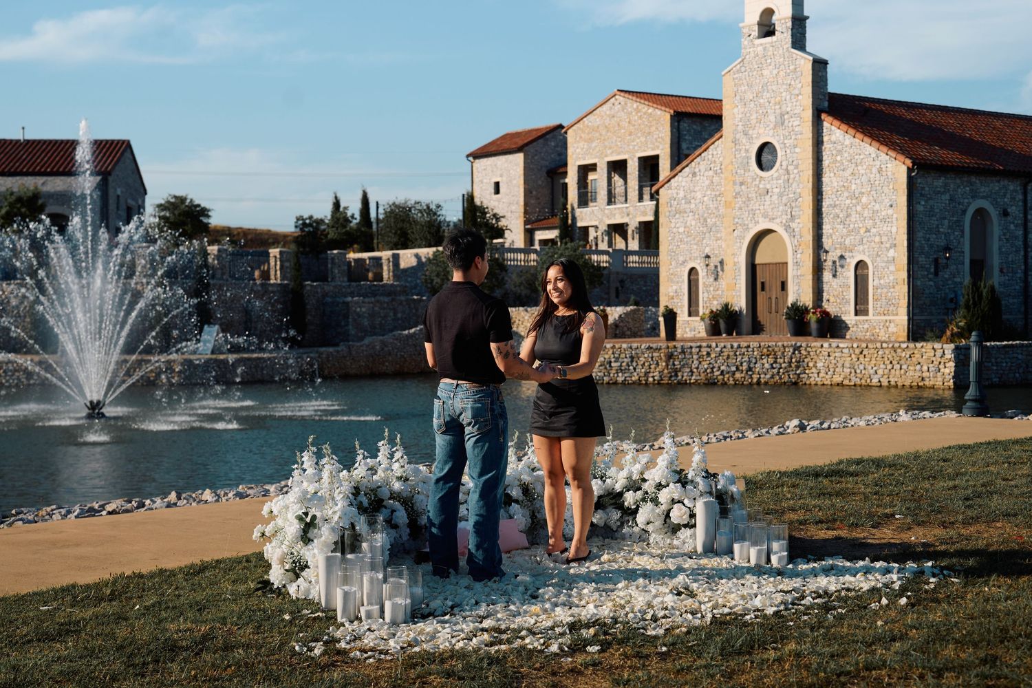 Couple sharing a romantic proposal moment by a fountain near a stone chapel with floral decorations.