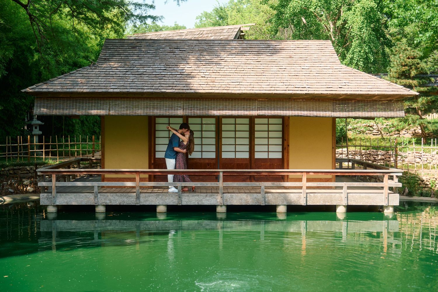 Couple embracing on the wooden deck of a traditional Japanese tea house beside a serene green pond.