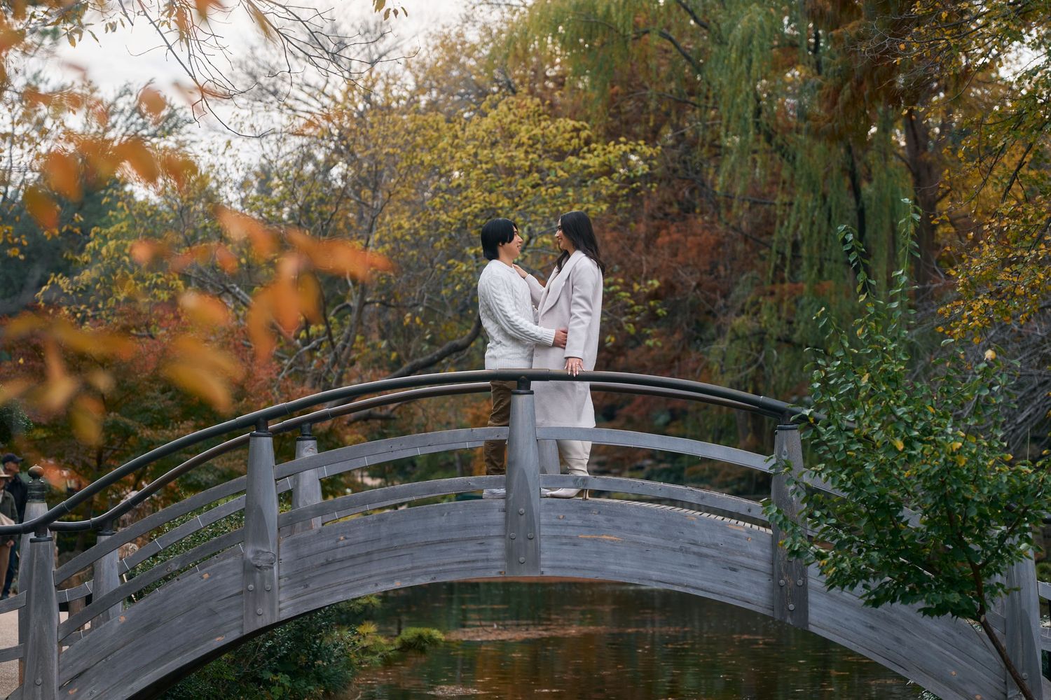 A couple in white clothing poses on a curved wooden bridge surrounded by autumn foliage in a garden setting.