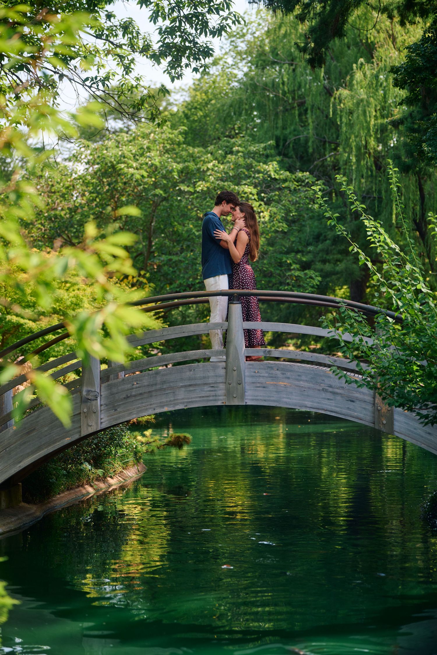 Couple embracing on a wooden arched bridge over a green pond surrounded by lush trees in a serene garden.