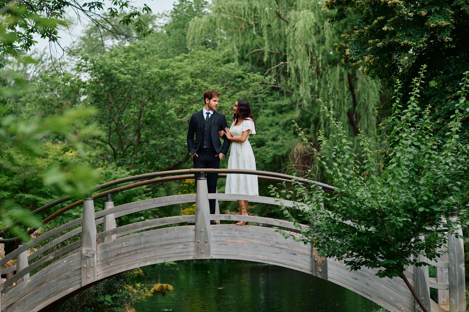 Couple standing on a wooden arched bridge surrounded by lush green trees in a romantic garden setting.