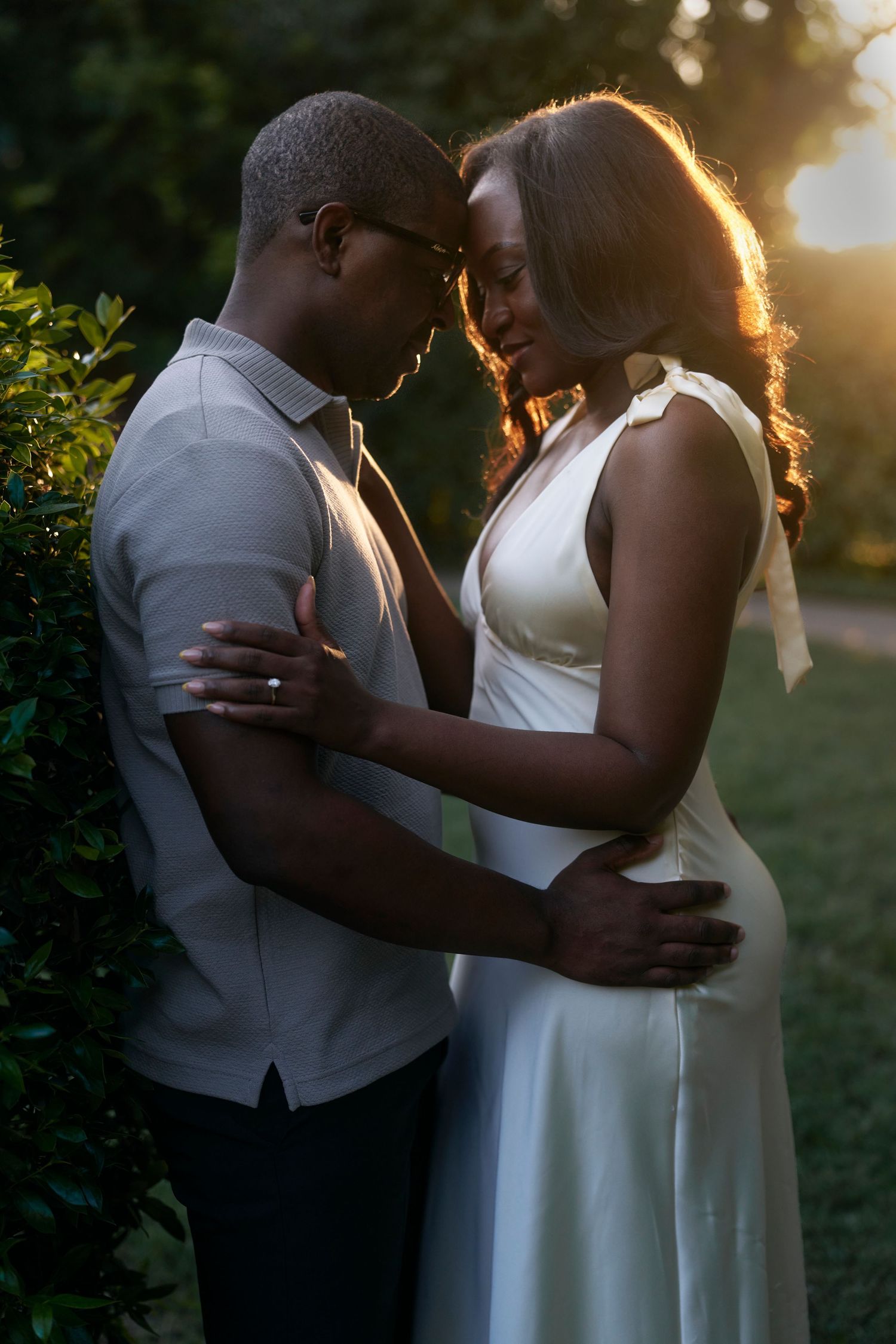 Romantic couple in white formal attire embracing during golden hour sunset in garden setting.