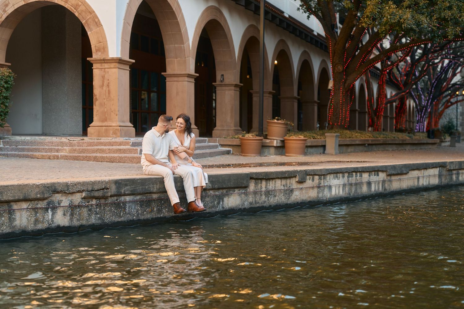 A couple sits romantically by a reflective pool under arched architecture in a sunlit outdoor setting.