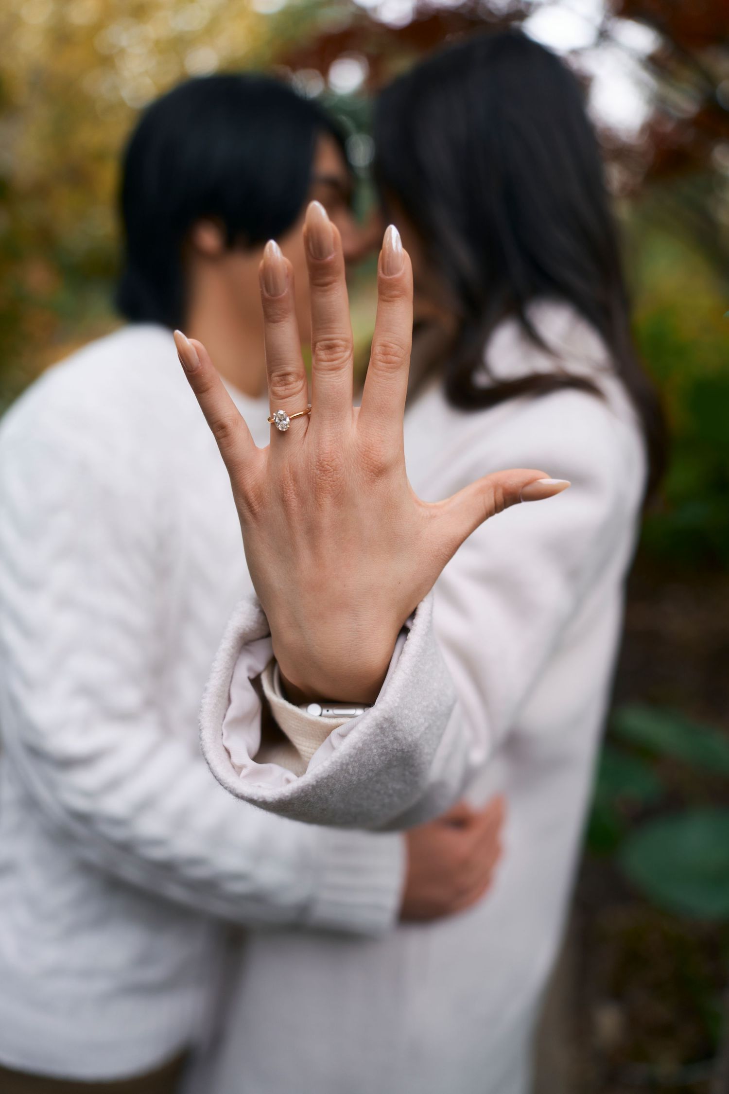 A close-up of an engagement ring on a hand with a blurred couple embracing in the background.