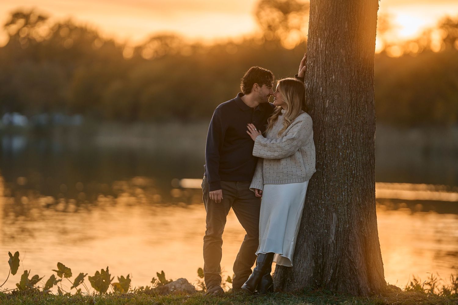 Intimate moment between couple leaning against tree during sunset by waterfront.