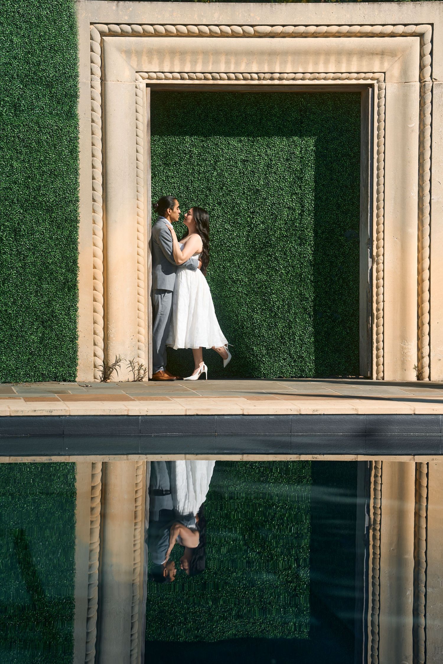 Couple embraces in elegant archway with green hedge wall and reflection in foreground.
