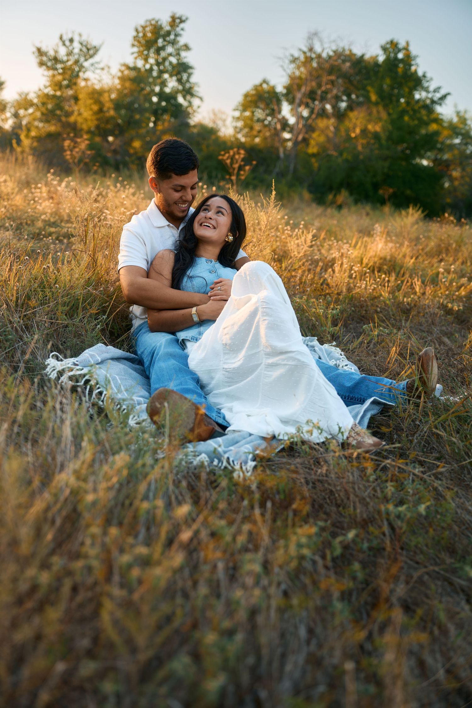 Couple in white and blue outfits lounging in golden grass during sunset photoshoot.