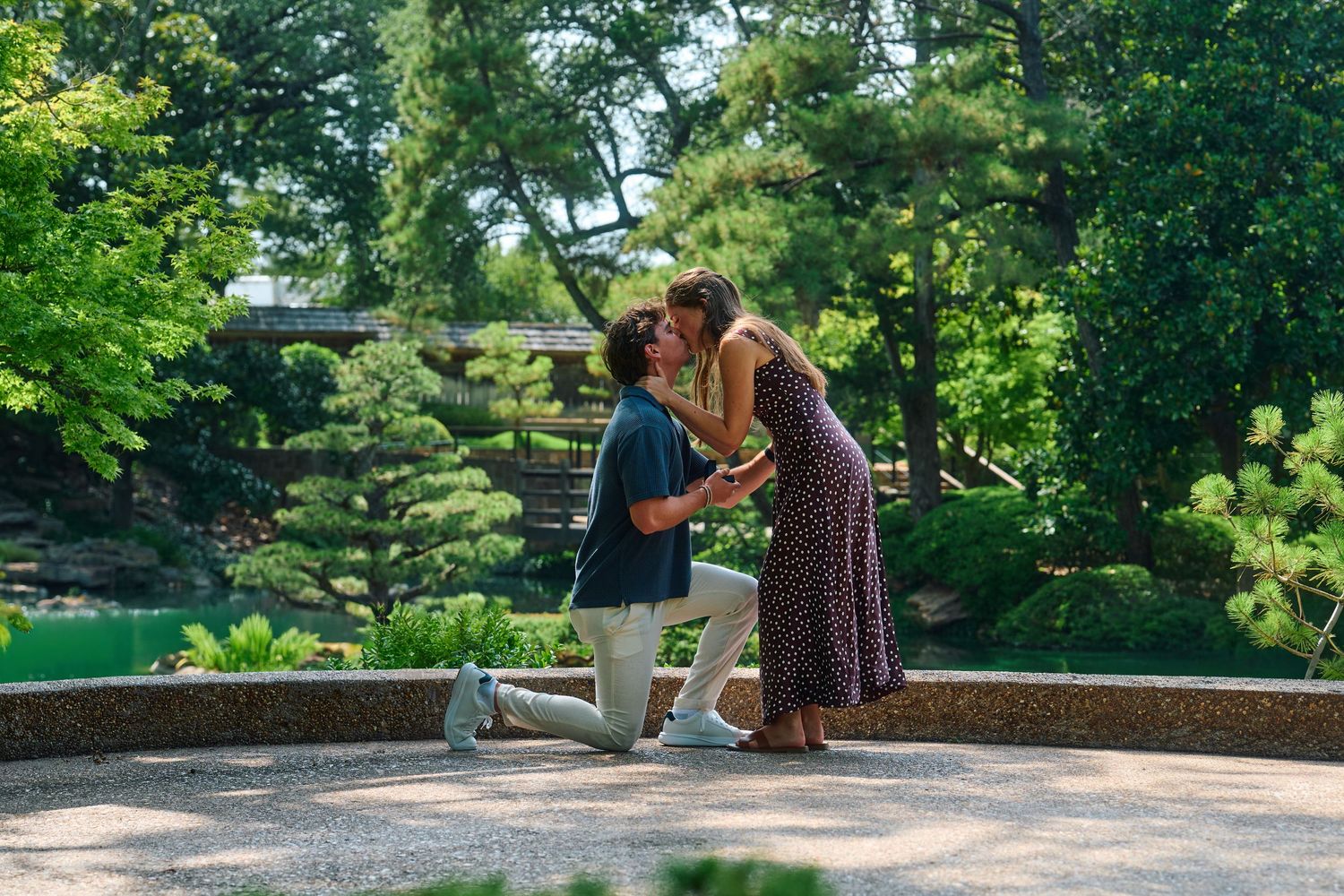 Man proposes to woman on one knee in a lush Japanese garden, sharing a romantic kiss near a serene green pond.