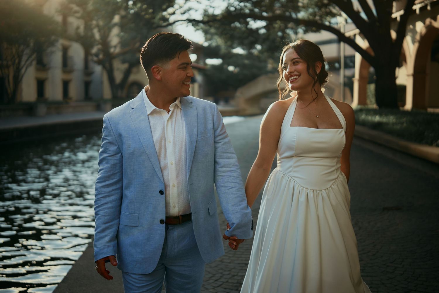 A couple in wedding attire walking hand in hand along a waterfront at sunset.