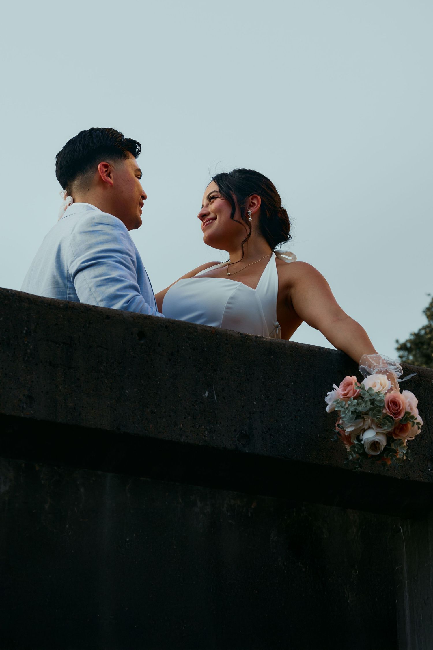Romantic silhouette of newlyweds embracing on a balcony with pink floral bouquet at sunset.