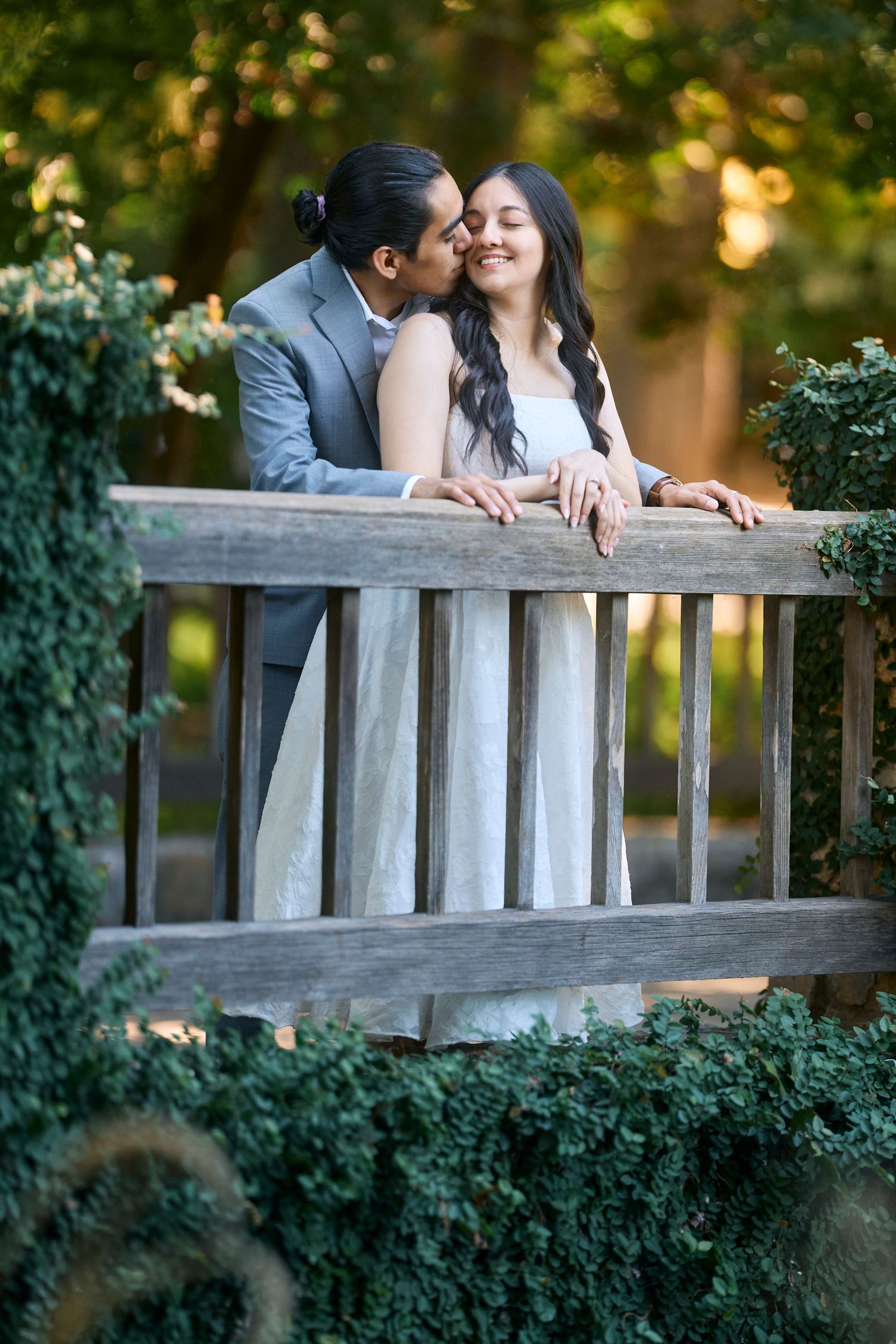 Romantic couple leaning against wooden fence surrounded by lush greenery.