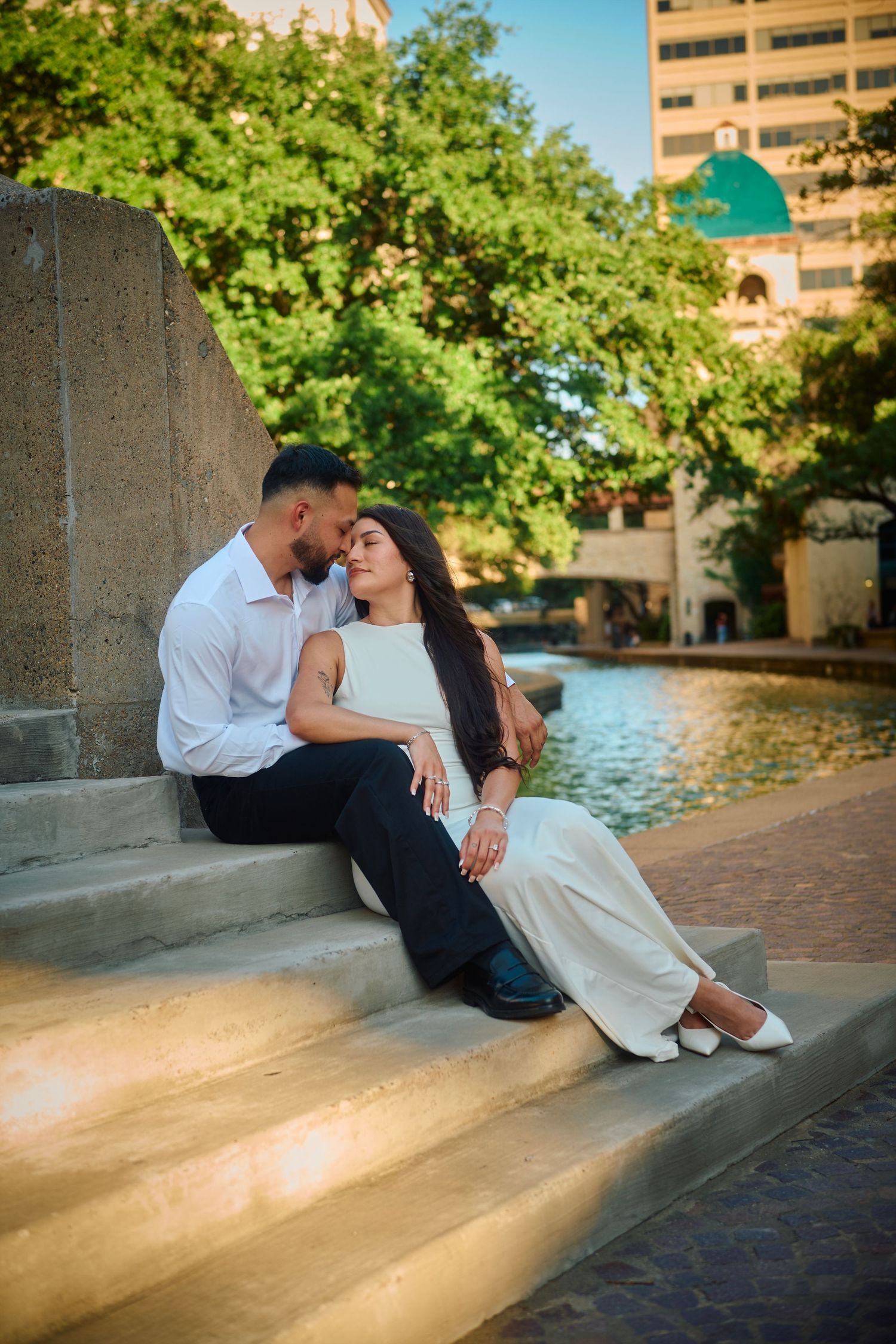Couple sitting on steps by a river walk, sharing a romantic moment in an urban park setting at golden hour.