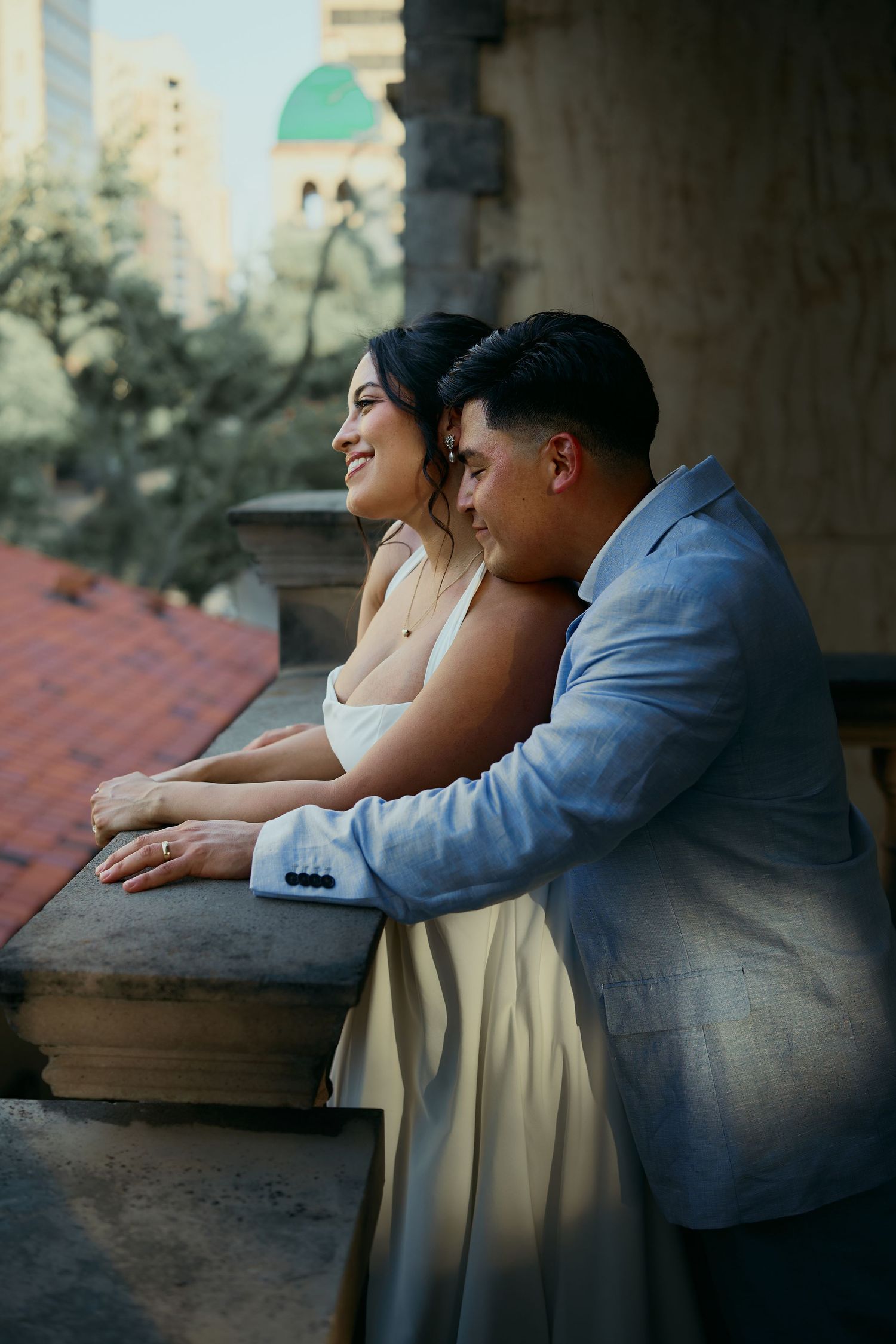 A couple in formal attire share a tender moment while leaning on a balcony railing at sunset.
