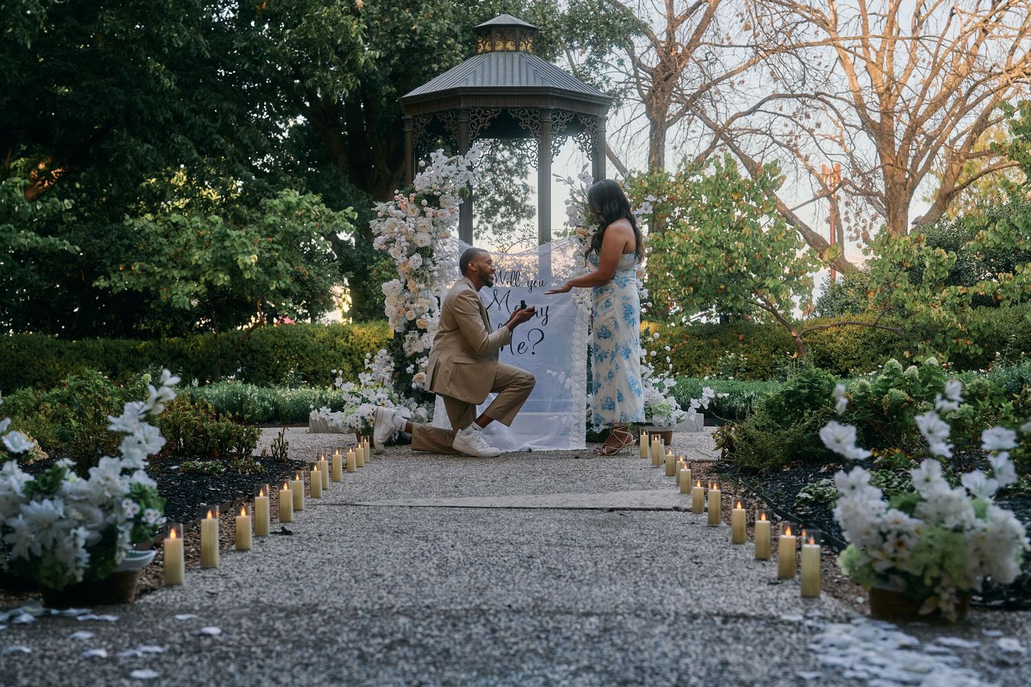 Man proposing to woman in elegant garden gazebo surrounded by candles and white floral arrangements at sunset.