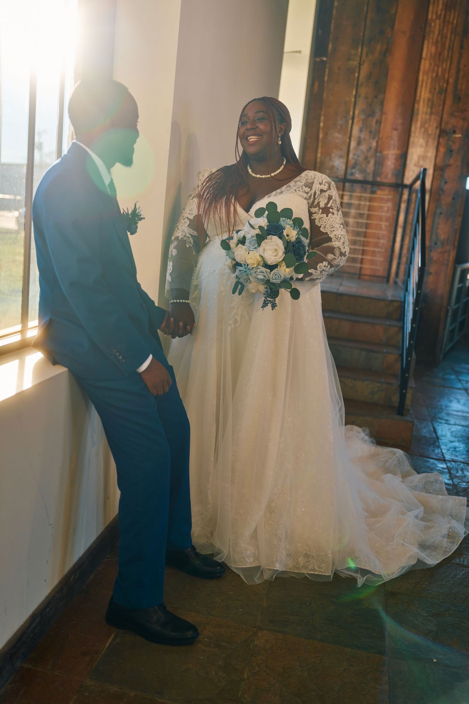 Smiling bride in lace wedding gown holding white bouquet stands with groom in blue suit by sunlit window.