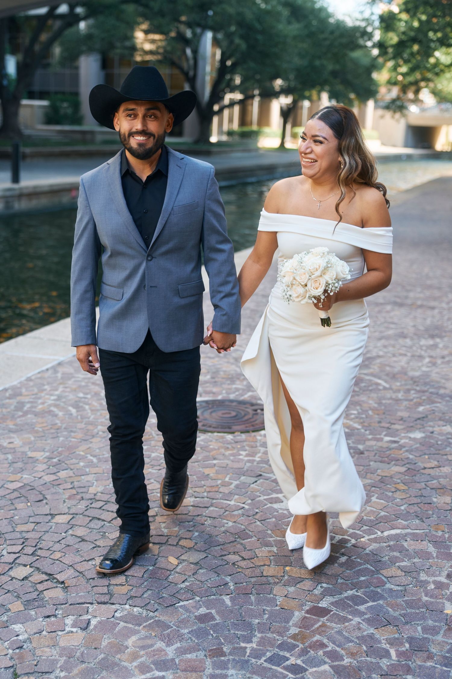 Bride and groom walking hand in hand on cobblestone path, groom in cowboy hat and gray blazer, bride in off-shoulder white gown.
