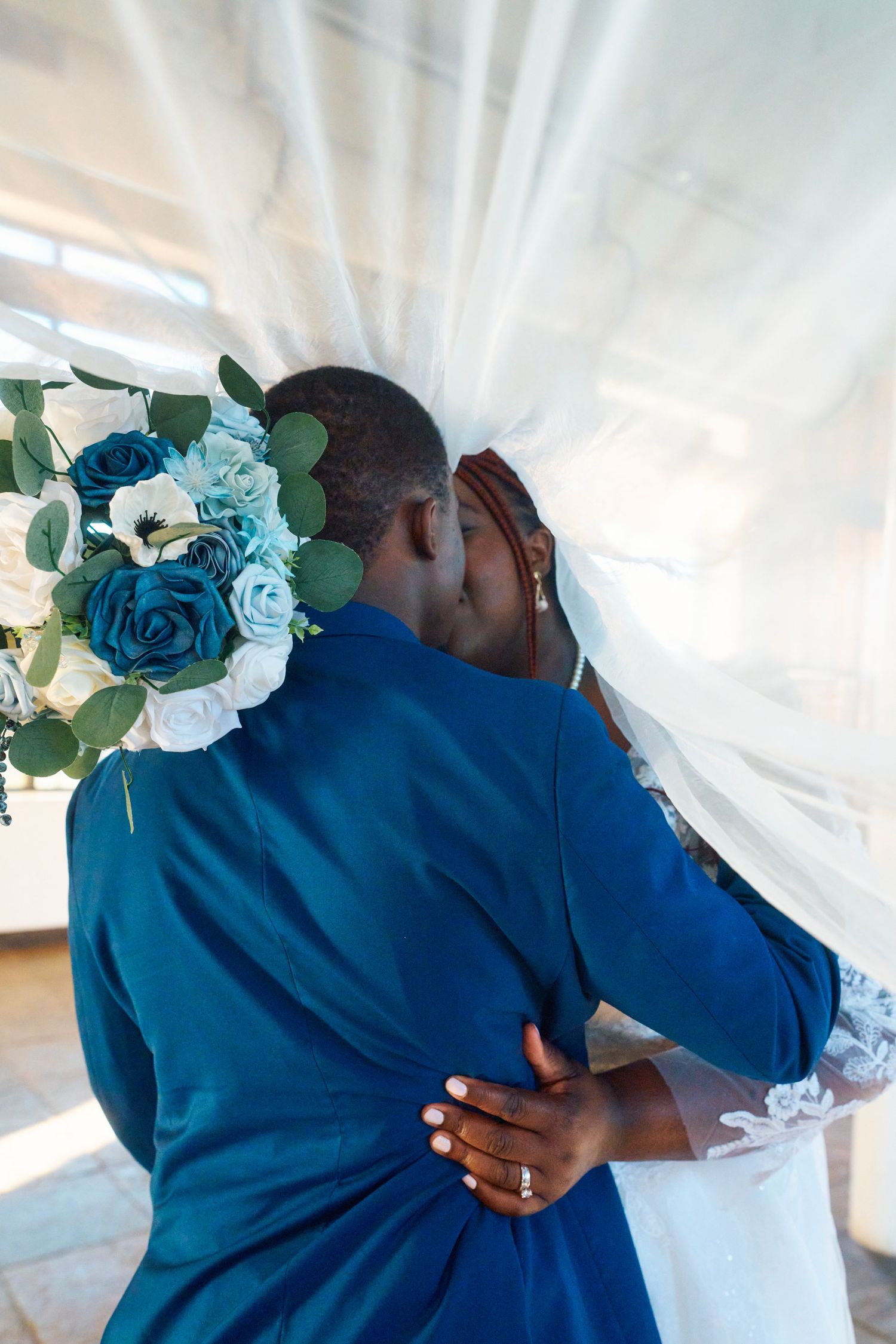 Two people in blue attire share an intimate embrace with teal roses and white curtains in the background.