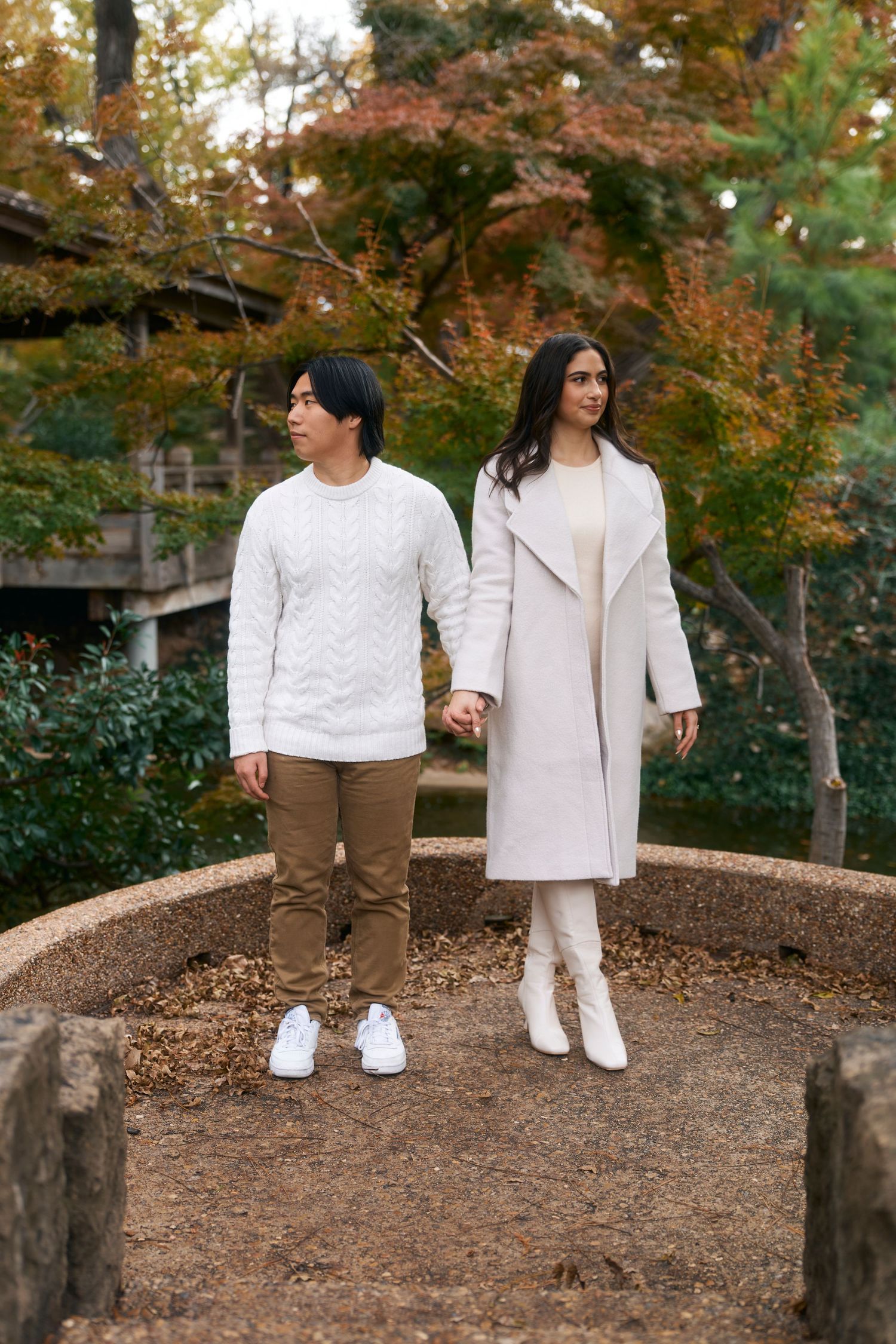 Two people in white outfits and brown pants walk together on a dirt path surrounded by autumn foliage.