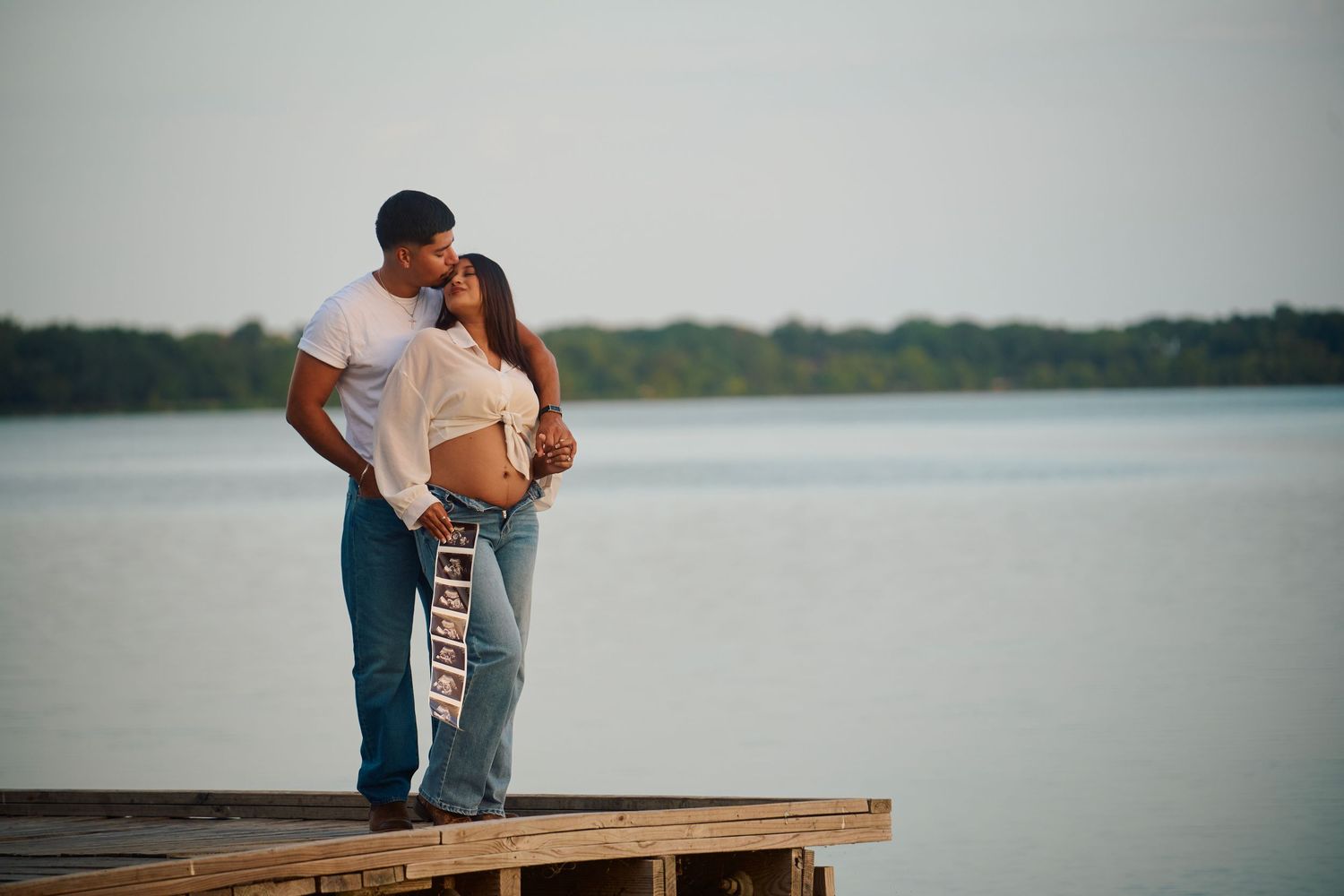 Couple sharing a tender moment on a wooden dock by a calm lake, with the woman showing a pregnant belly.