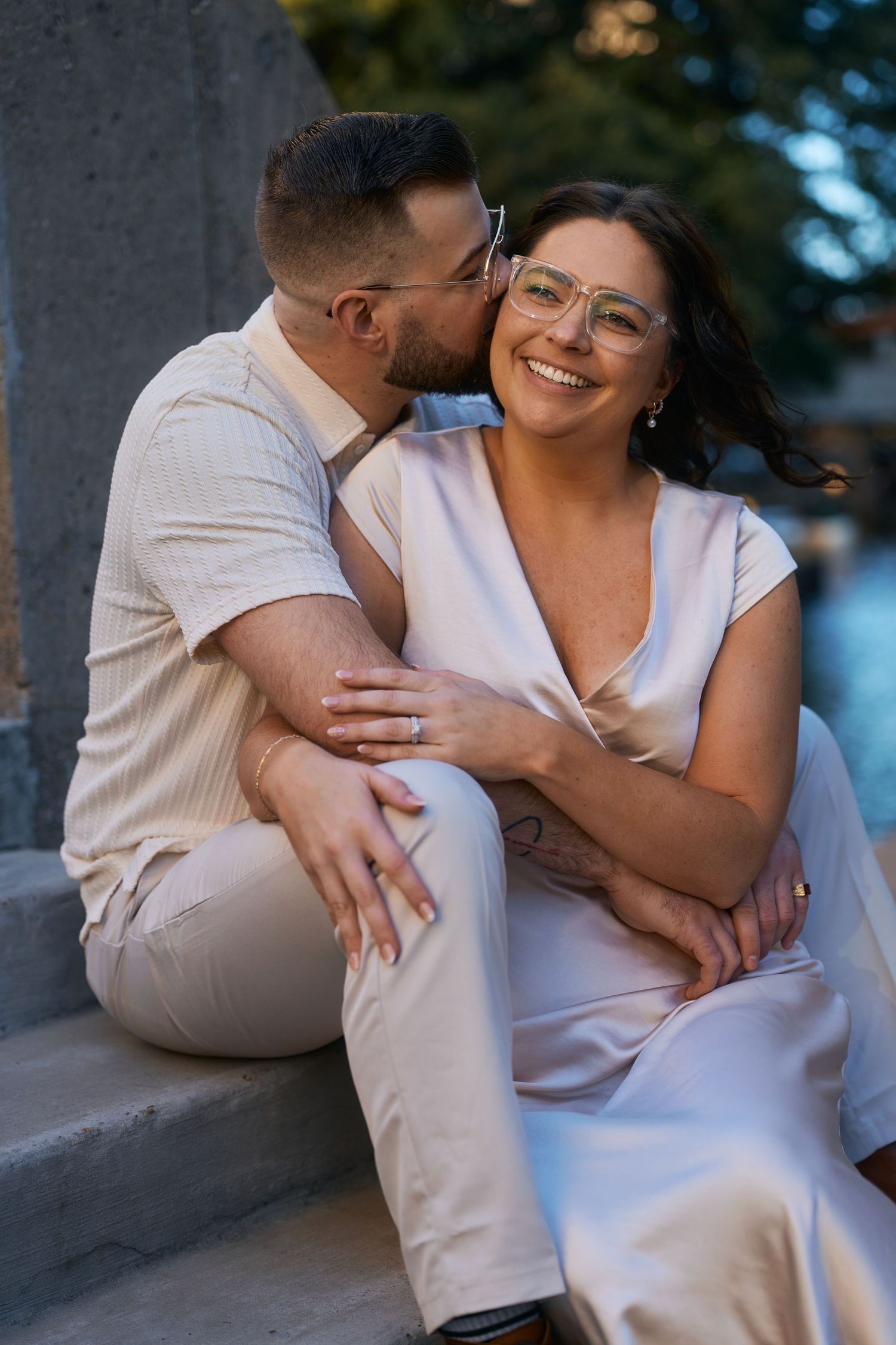 Couple sitting on stone steps, man kissing woman's cheek, both wearing beige outfits, woman smiling and wearing glasses.