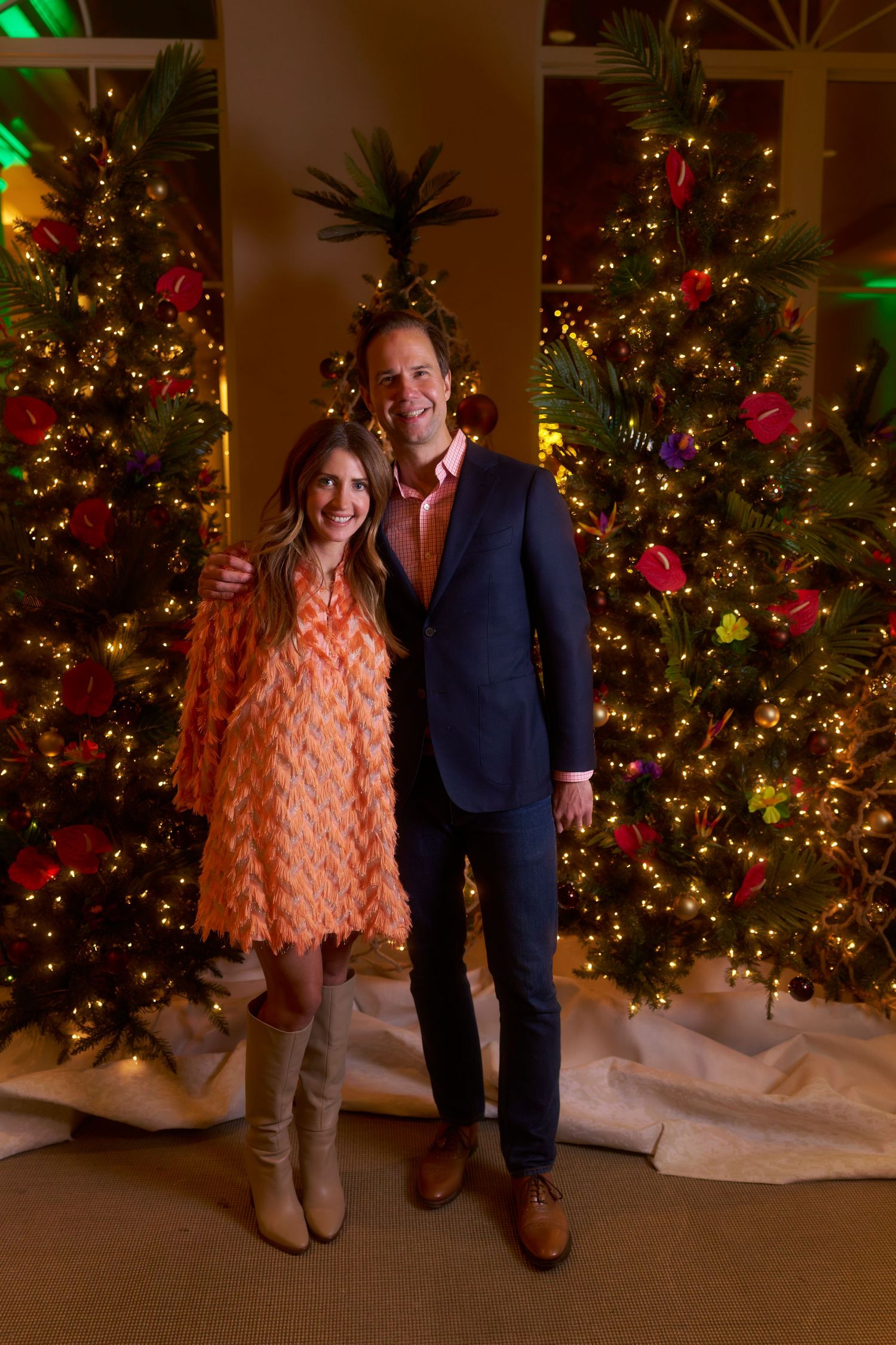 A couple in festive attire poses in front of illuminated Christmas trees at a holiday event.