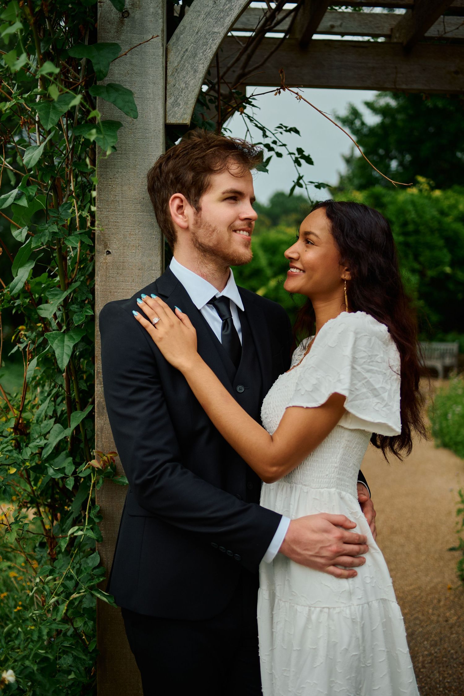 A couple in formal attire embraces outdoors under a garden pergola, smiling at each other romantically.