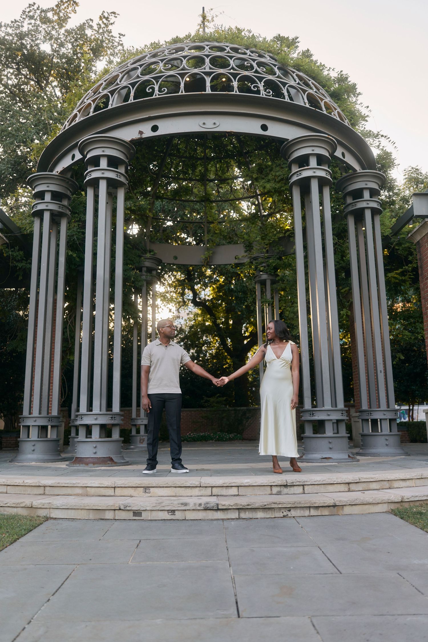 Couple holding hands under an ornate metal gazebo with domed roof in a garden setting.